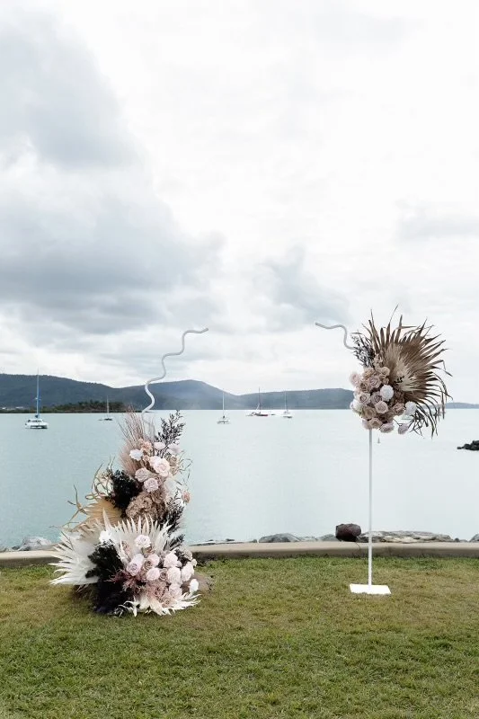 Decorative floral arrangements on a grassy area by a body of water with sailboats and hills in the background, suitable for an outdoor event or wedding.