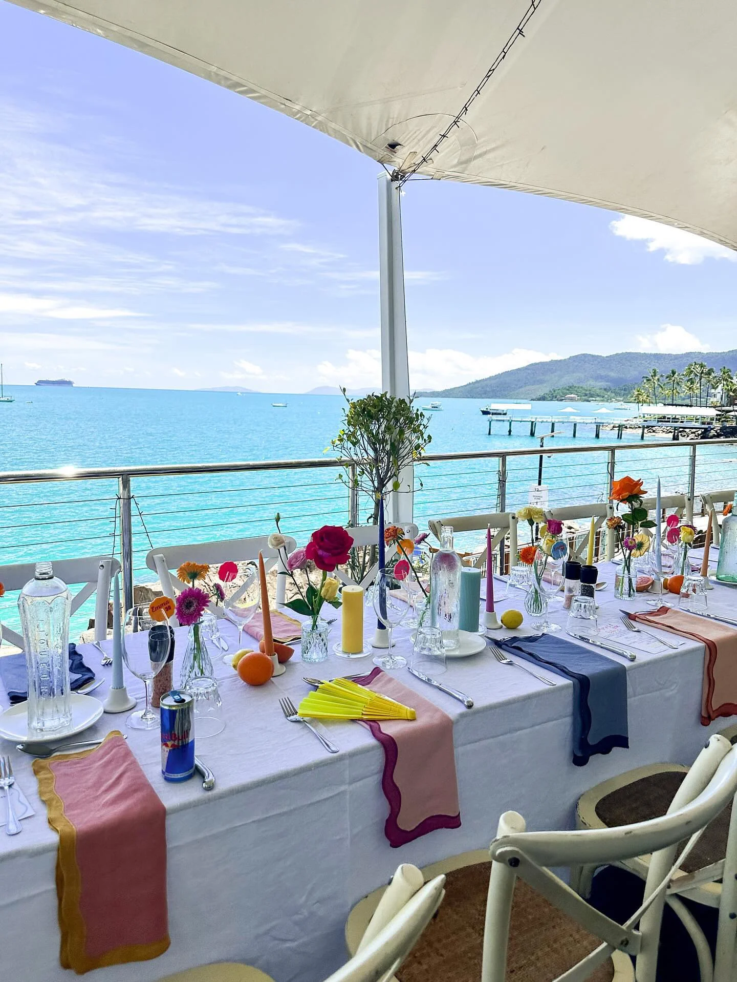 A seaside outdoor dining setup with a long white tablecloth-covered table decorated with colorful flowers in small vases, candles, and tableware, overlooking turquoise water, with boats and a mountain in the background under a canopy.