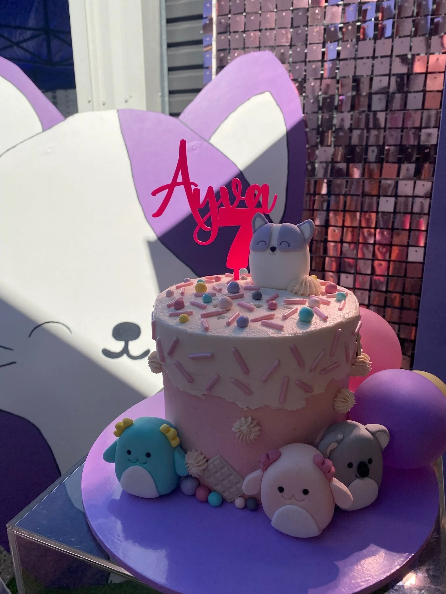 A pink birthday cake with a white cat topper, colorful sprinkles, and fondant animal decorations around it. A pink birthday sign with the name 'Alya' and number '7' is on top, with the background featuring a large cartoon-style cat with a purple and 