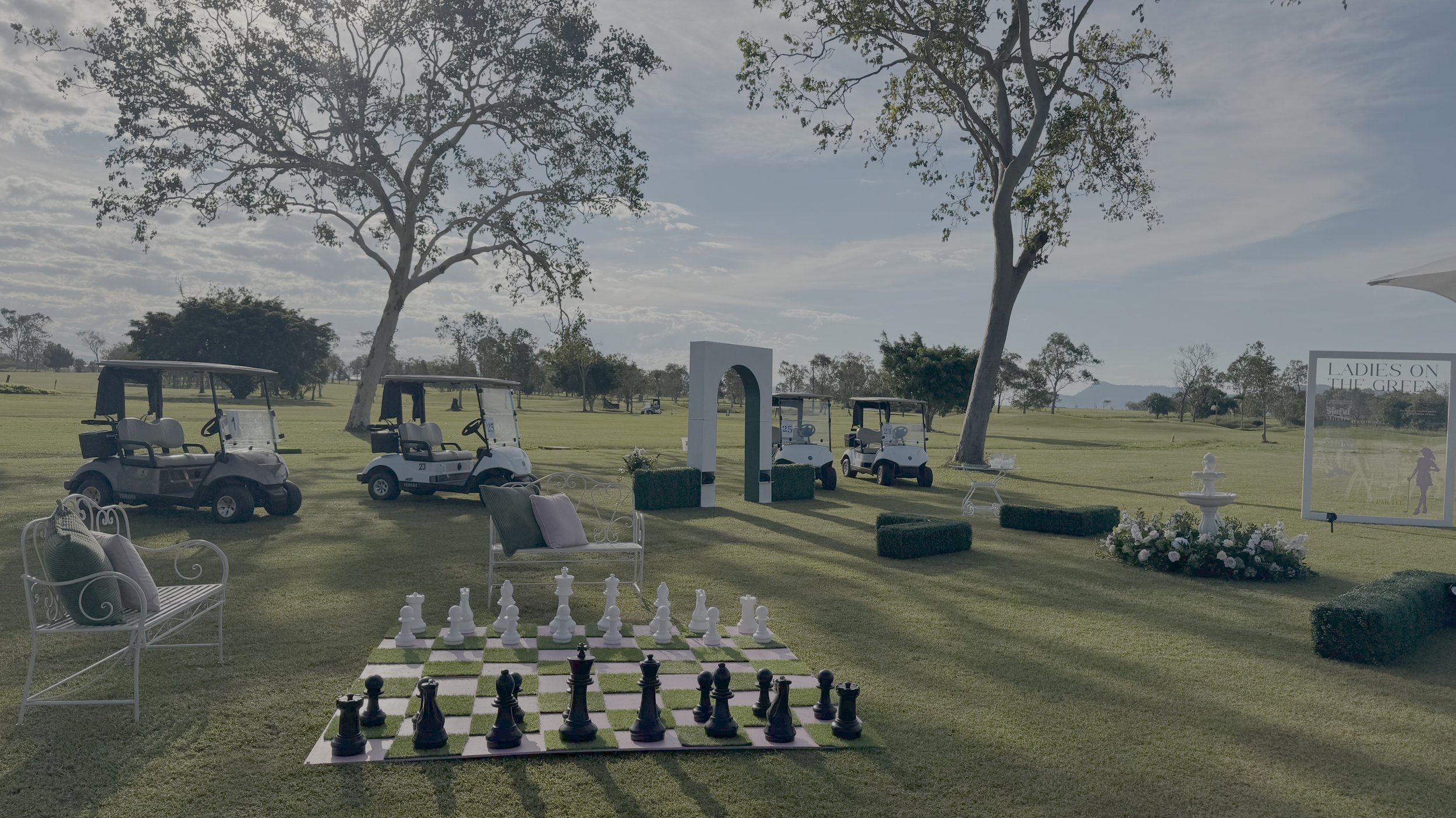 A scenic outdoor setting at a golf course with four golf carts parked, a large chessboard with pieces in the foreground, decorative benches with cushions, a white archway, a cluster of flowering plants, a fountain, and a sign that reads "Ladies on th