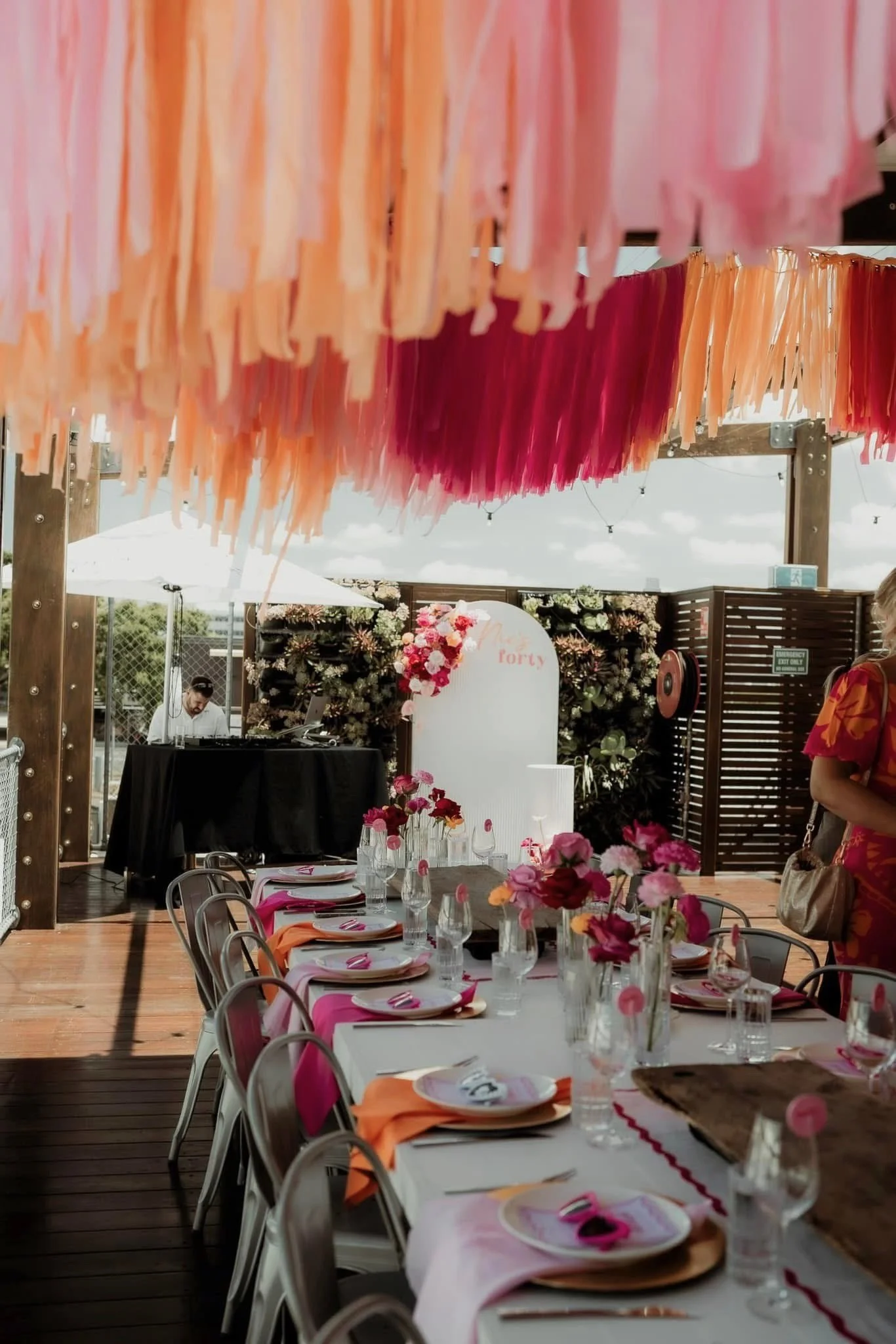 Decorated outdoor event space with pink and orange paper tassel banners hanging from the ceiling, a long table with pink, orange, and white napkins, floral centerpieces, and a backdrop with flowers and the word 'forty' in pink lettering.