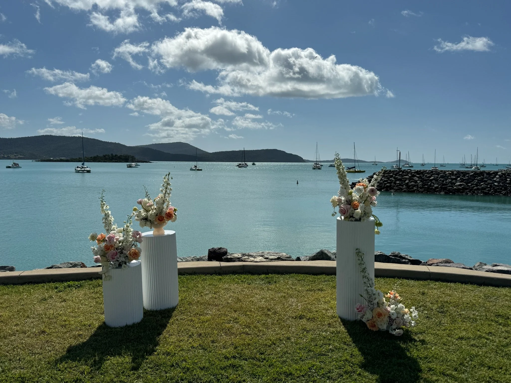 Outdoor wedding setup with two white flower arrangements on tall white pedestals near a waterfront, sailing boats anchored in the water, mountains in the background, under a partly cloudy sky.