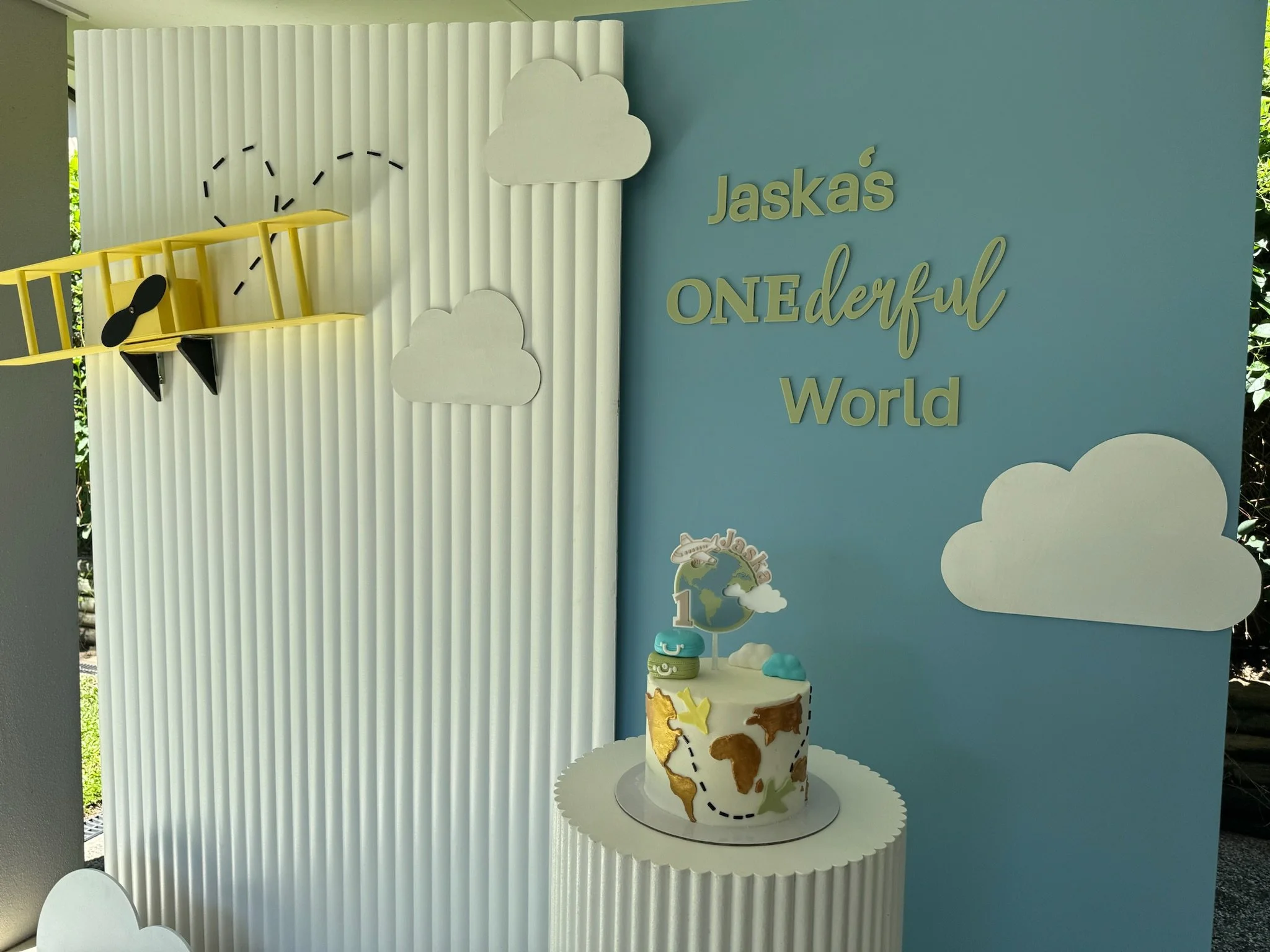Decorative display for a child's birthday with a cake featuring a world map, clouds, and an airplane, and a backdrop with the message "Jaska's ONEderful World."