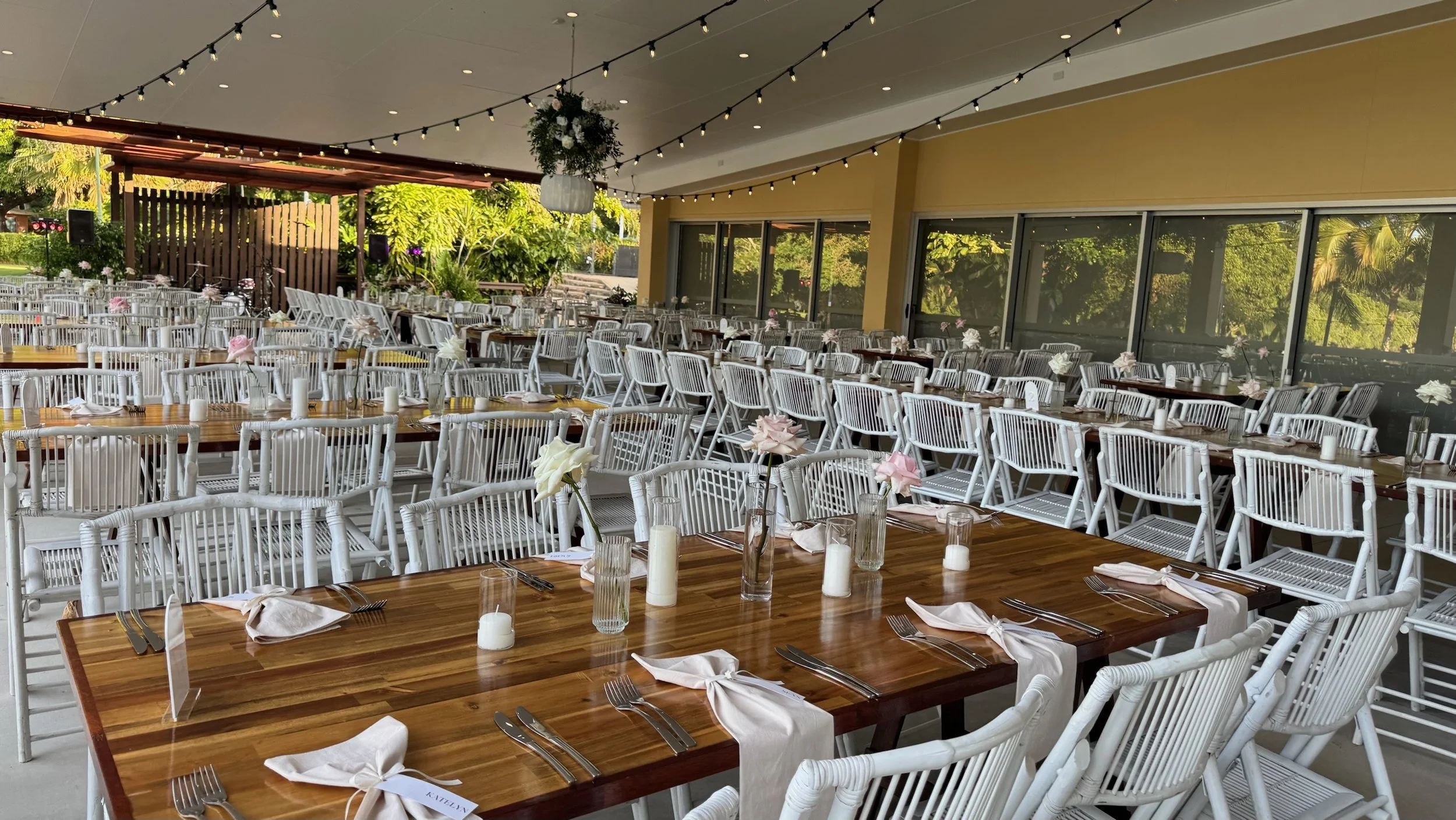 Wedding reception setup with wooden tables, white chairs, floral centerpieces, candles, and string lights inside a glass-enclosed patio.