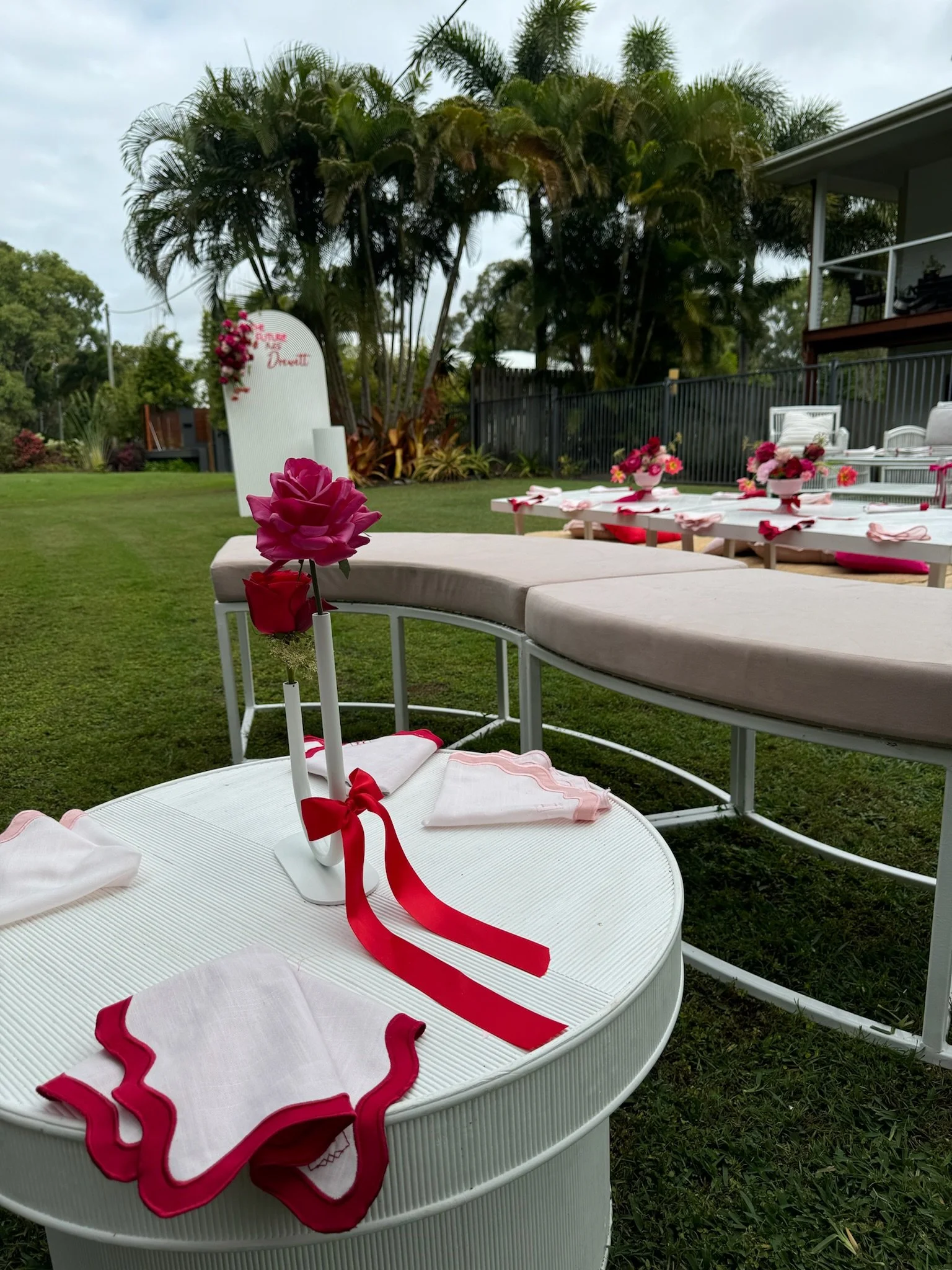 Outdoor setup with white round table, pink floral centerpiece, white napkins with pink borders, and a curved bench, decorated for a celebration with pink flowers and a sign in the background.