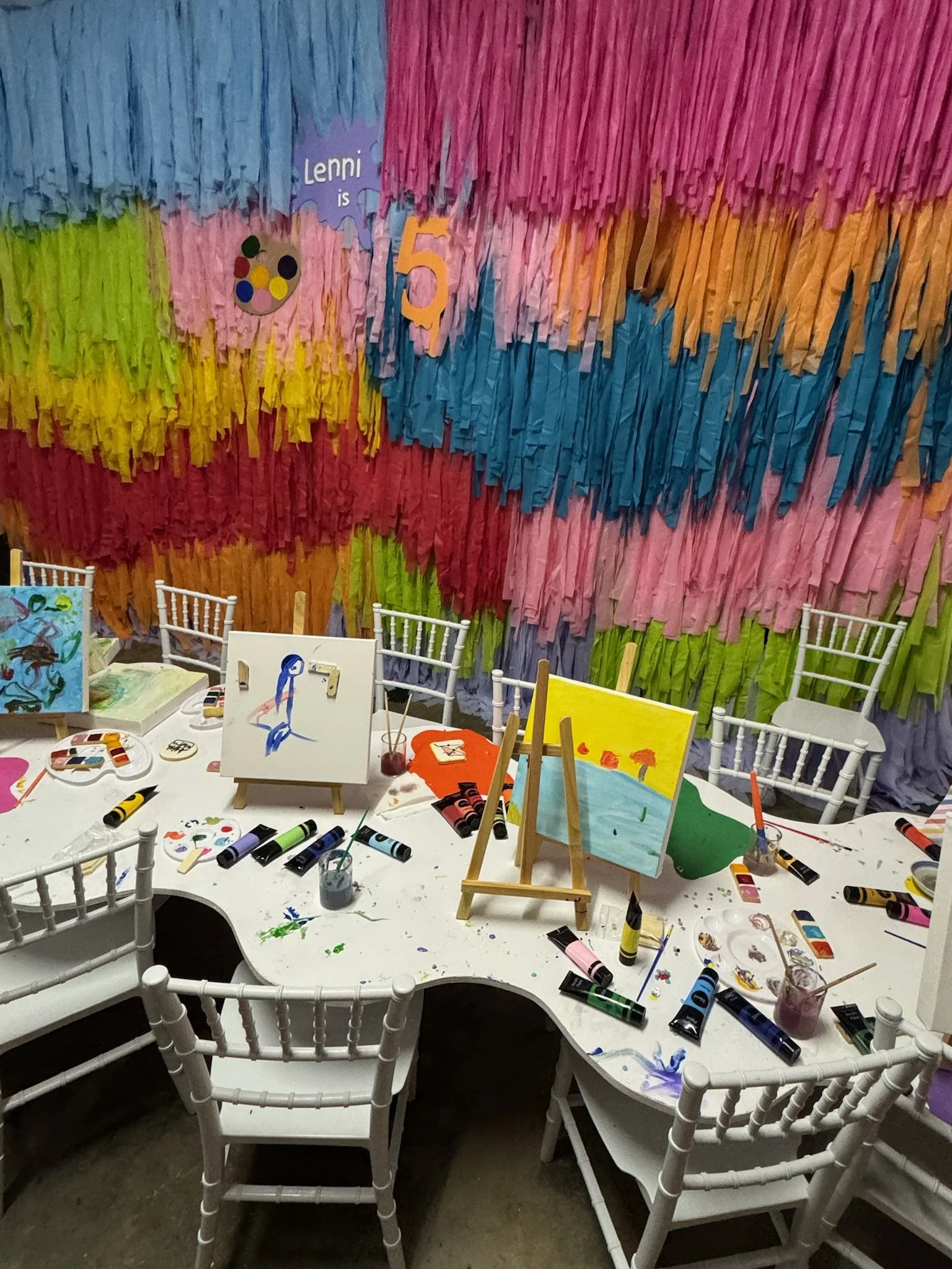 Colorful birthday party decoration with a rainbow tissue paper backdrop, white table covered with paint supplies, small canvas paintings, and white chairs.