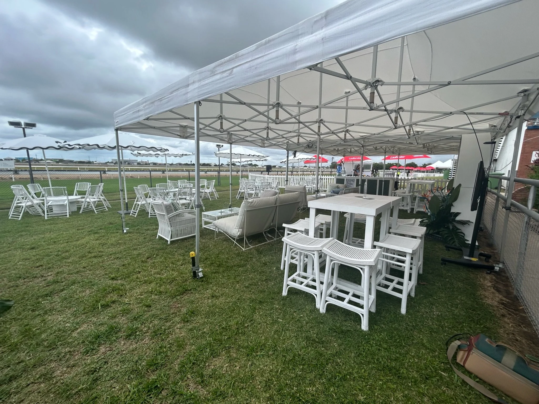 Outdoor event setup with white canopy, white tables, white chairs, and lounge seating on a grassy field under cloudy sky.