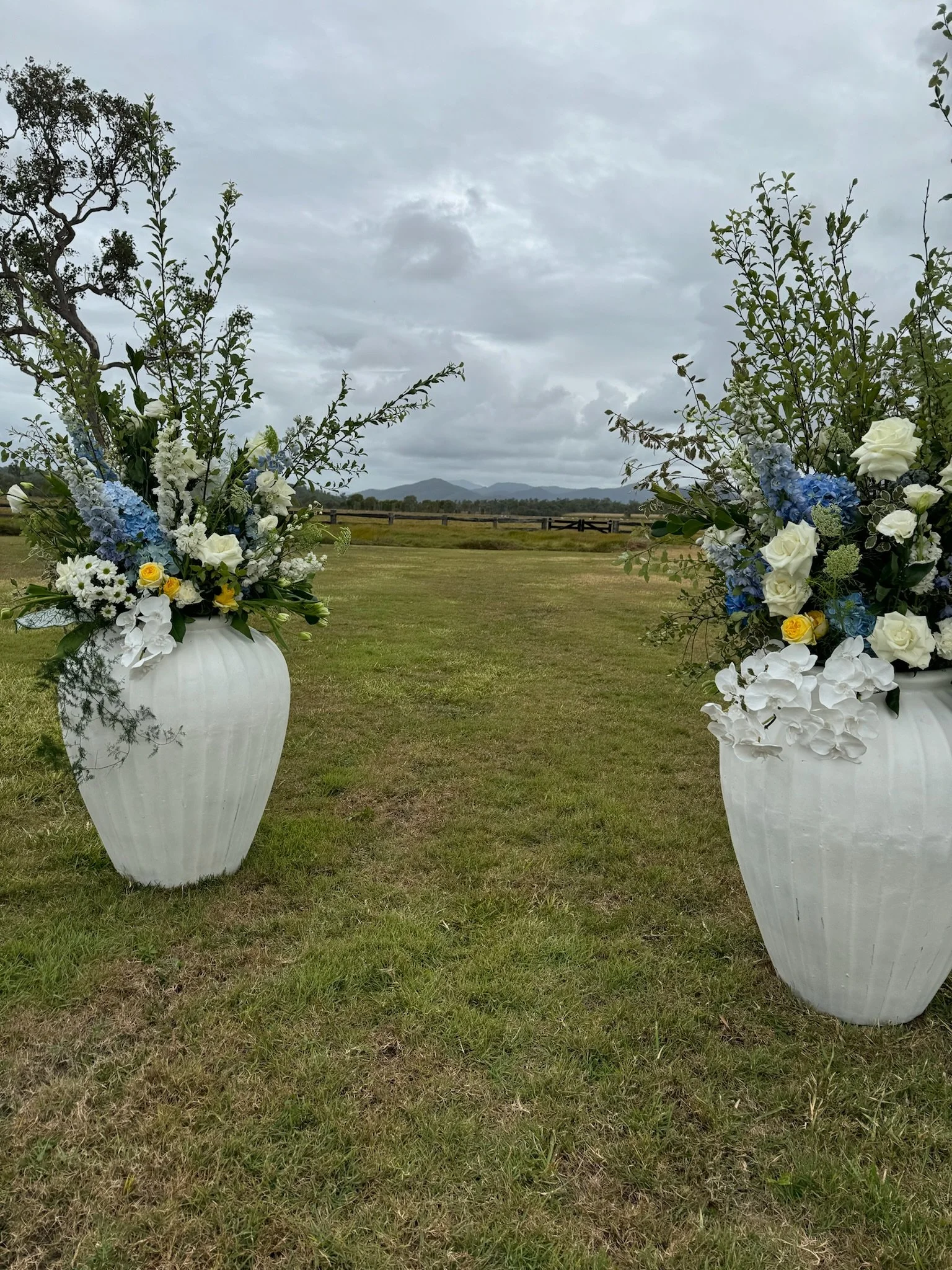 Two large white vases filled with blue and white flowers, green foliage, and small yellow flowers, placed on a grassy field with a cloudy sky and distant mountains in the background.