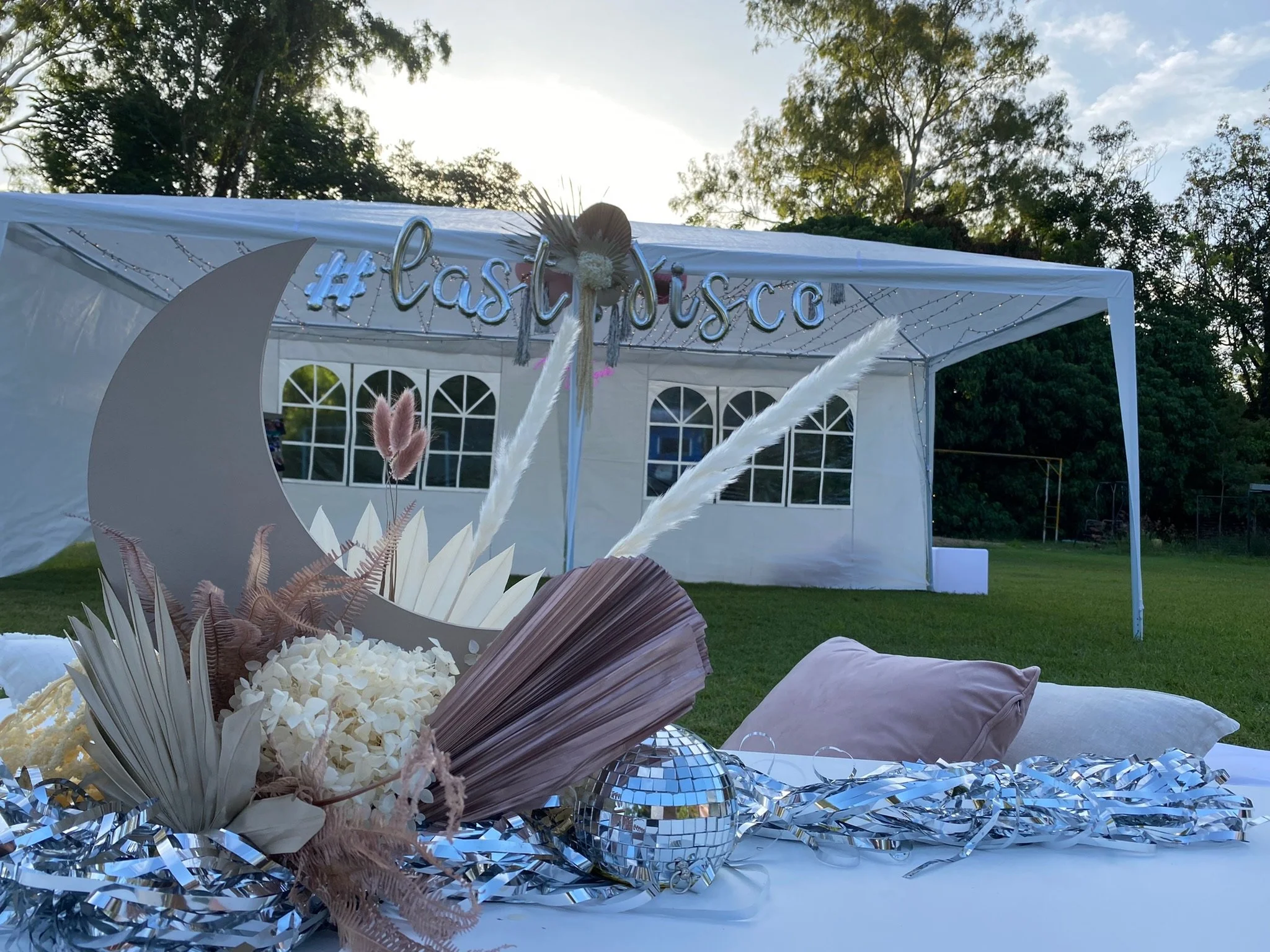 Decorated outdoor tent with a '#lastdisco' sign, featuring a table with floral arrangements, silver tinsel, and disco ball, set in a grassy area with trees and a partly cloudy sky.