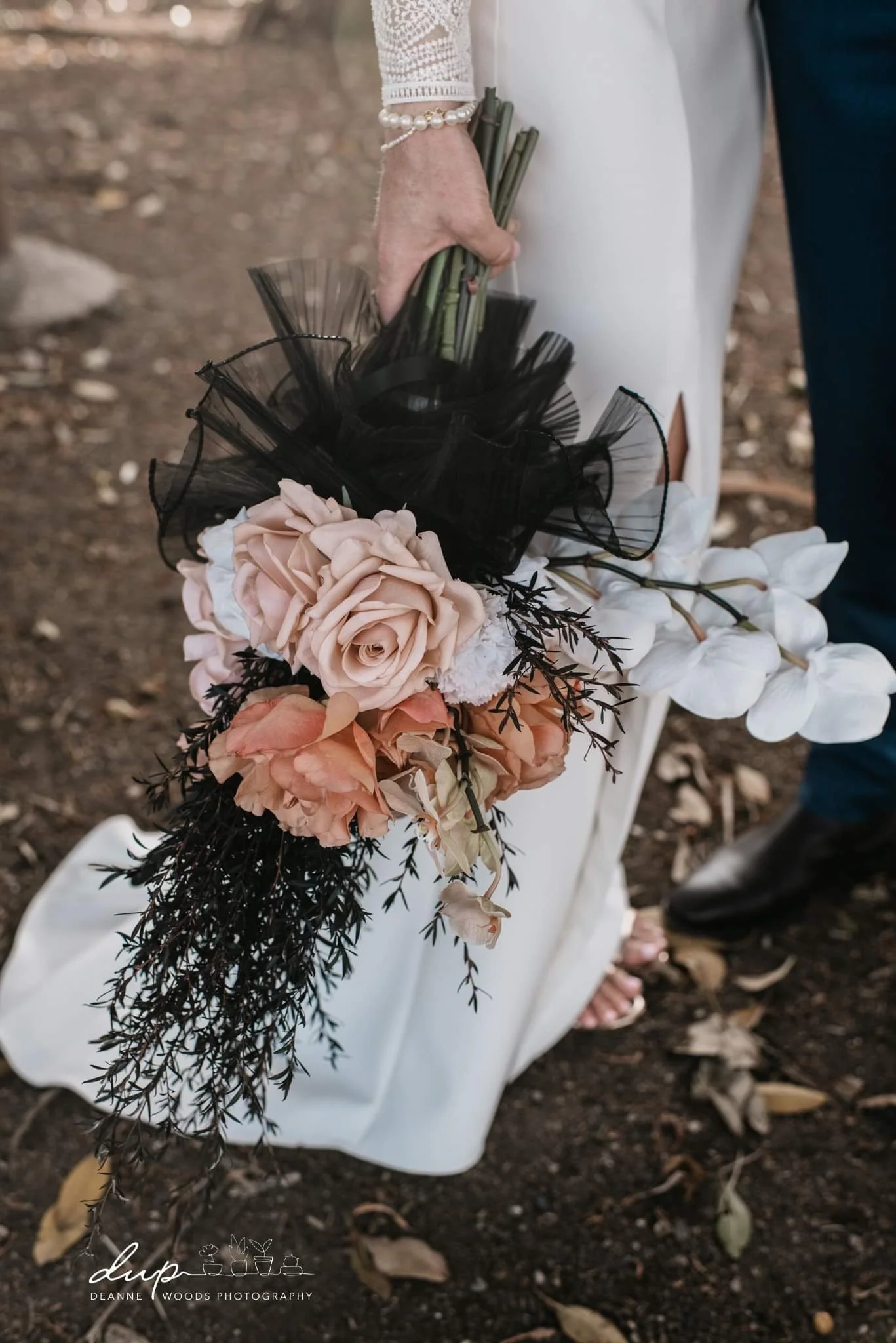 A bride holding a bouquet of pink and white roses, white orchids, dark foliage, and black mesh decoration, with both individuals dressed in wedding attire.
