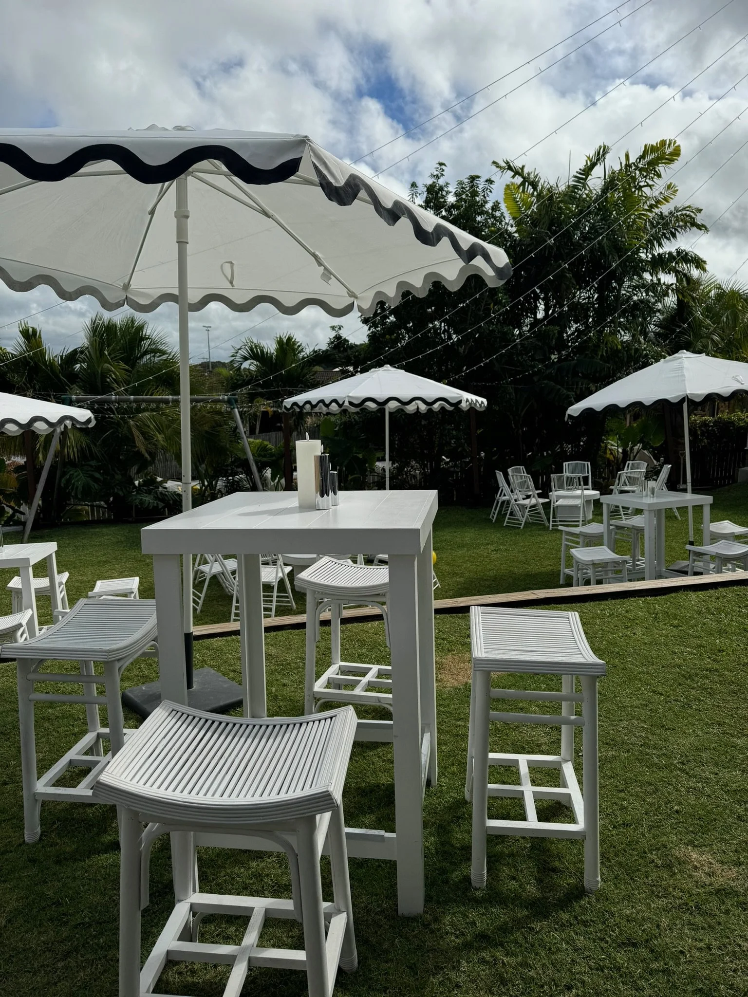 Outdoor patio area with white tables, chairs, and umbrellas on a grassy lawn, with trees and overcast sky in the background.