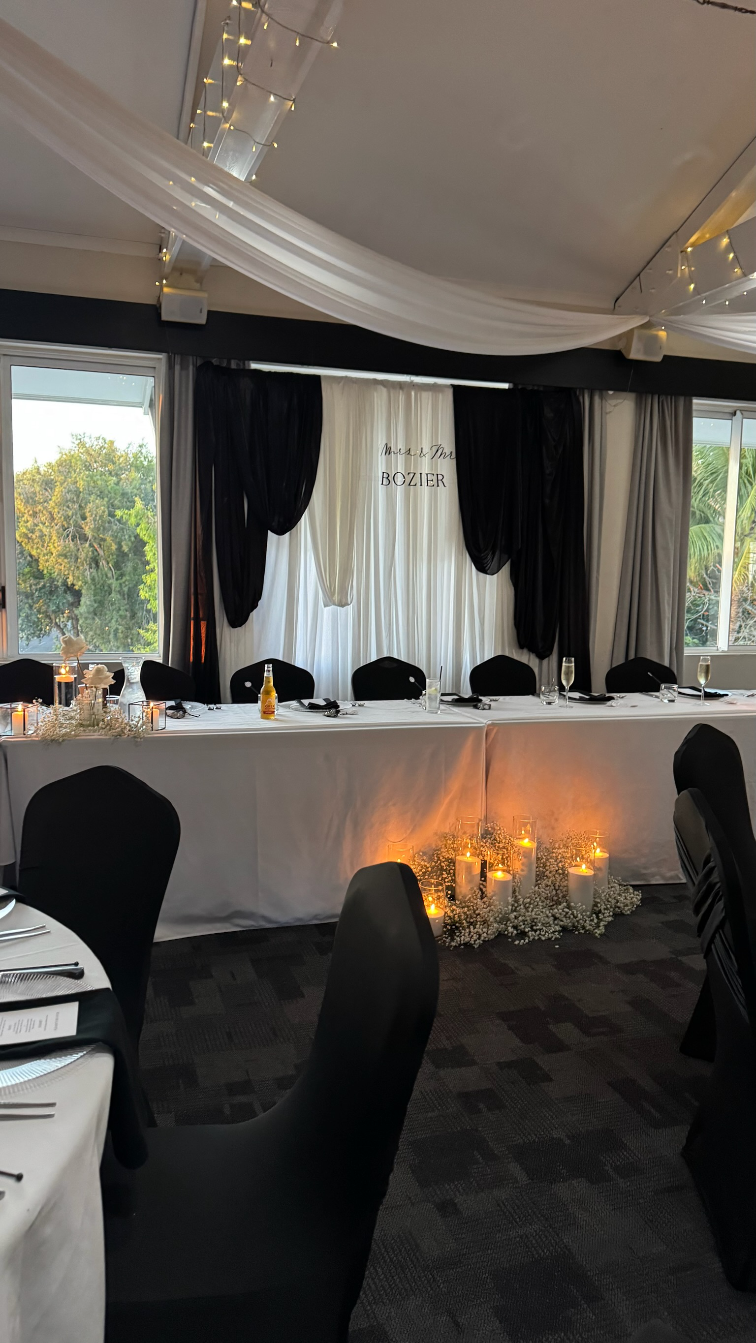 Elegant wedding reception setup with a long bridal table decorated with candles and flowers, black and white drapery backdrop with 'Mrs. & Mr. Bozier' written on it, and tables with place settings and glasses, with windows showing greenery outside.