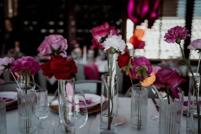 Table decorated with pink, red, white, purple, and orange flowers in tall glass vases at a social event or celebration.