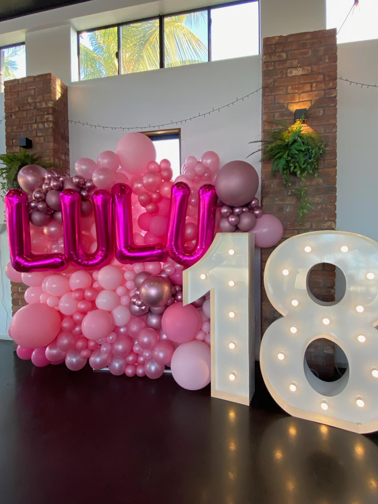 Decorative balloon arrangement with pink, purple, and metallic balloons, forming the word 'LULU' in metallic pink balloons, and a large illuminated number '8' on a dark floor, against a brick wall with plants and windows
