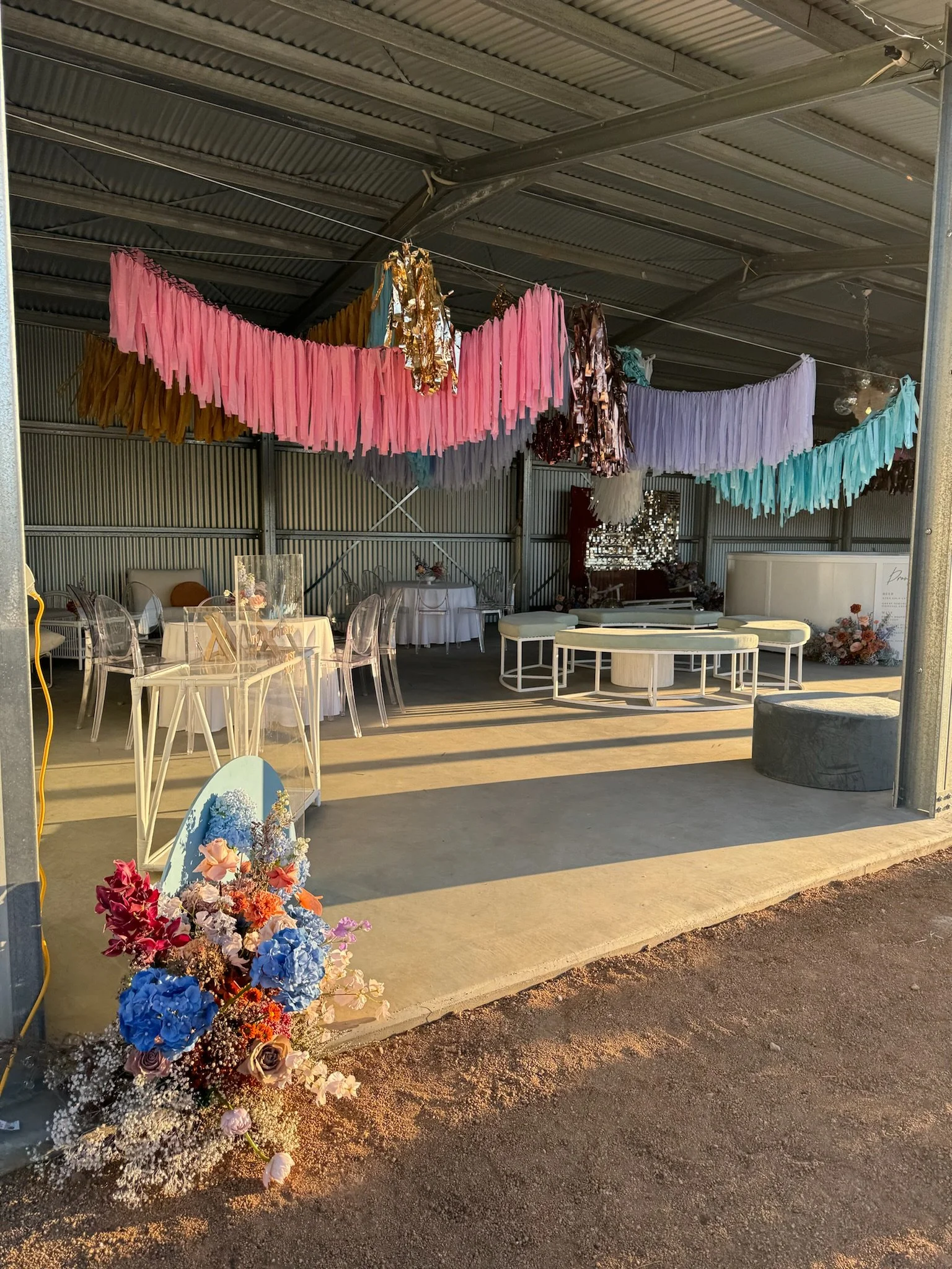 Party or event setup with colorful streamers, floral arrangements, transparent and white chairs, and decorative furniture inside a metal barn-like structure.
