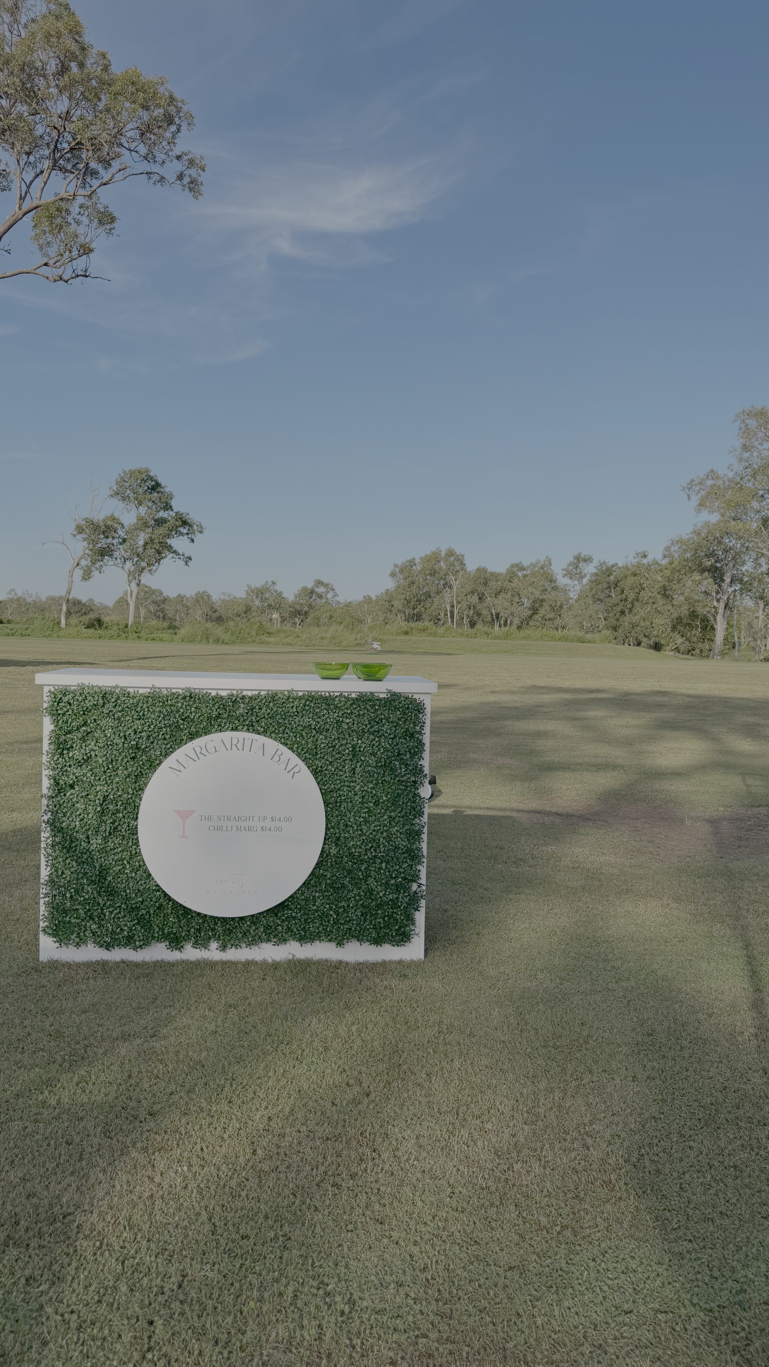 An outdoor scene with a white rectangular structure covered in green foliage, holding a round white sign with text. Two green bowls are on top of the structure. The area is grassy with trees in the background and a clear blue sky overhead.