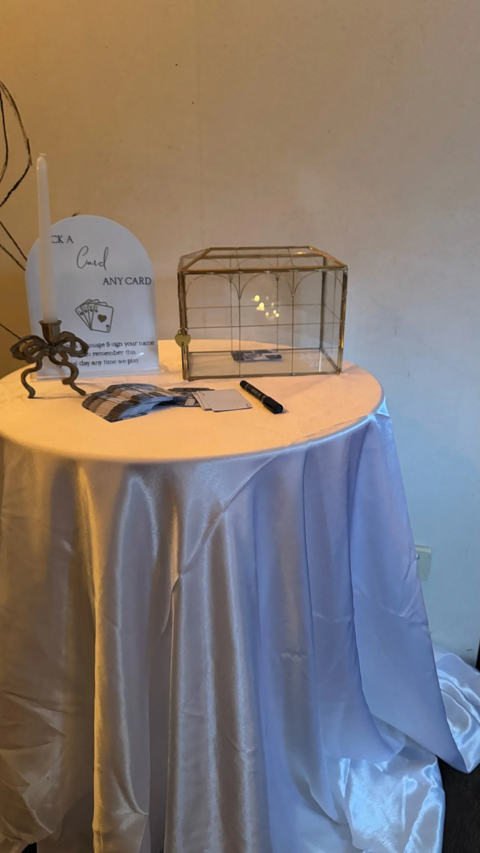 A small round table covered with a satin tablecloth displaying various items, including a white candle holder with a candle, a paper sign, a glass display case, a black marker, and a few stacks of cards.