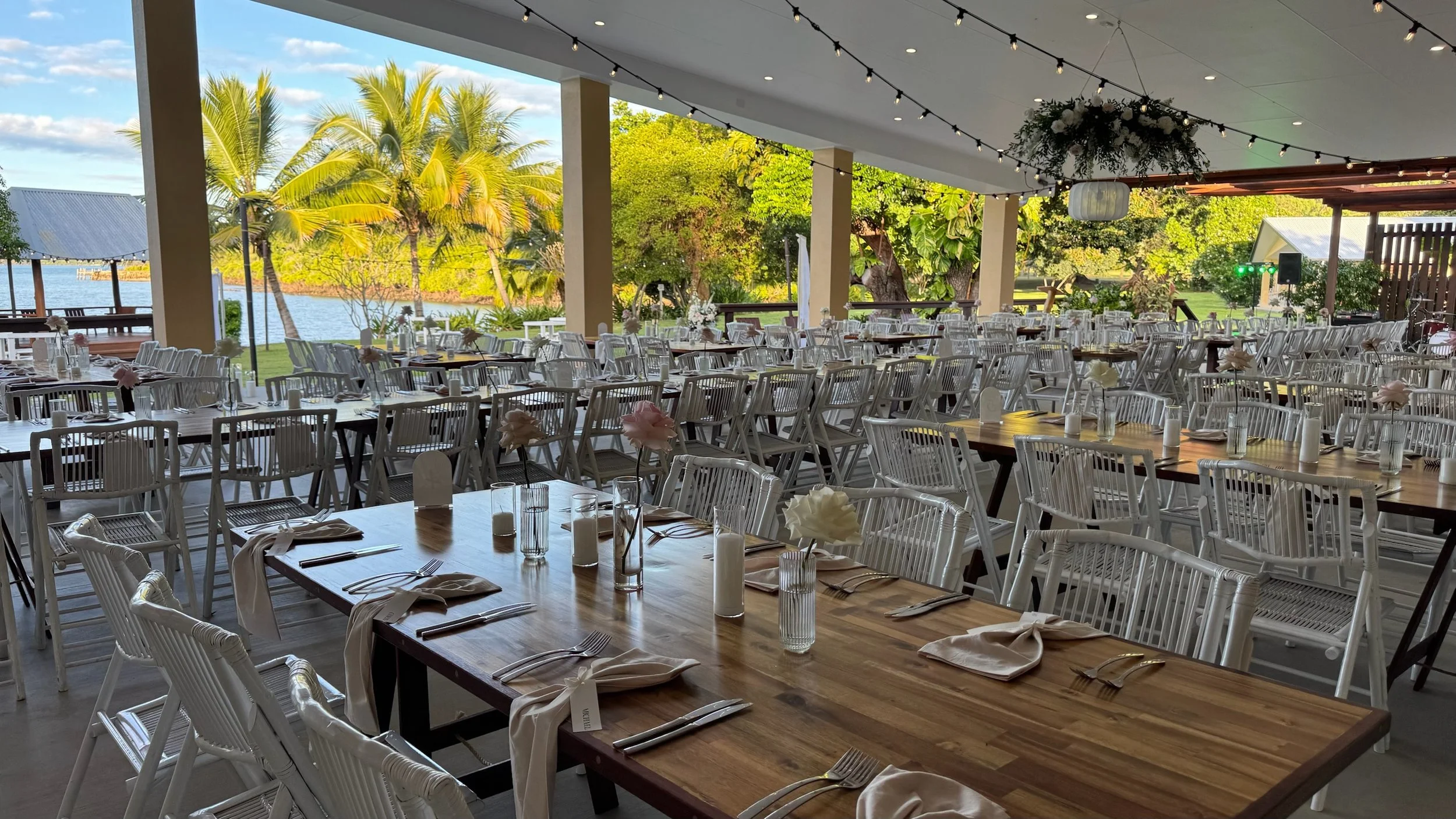 Indoor banquet setup with wooden tables, white chairs, and elegant table settings, overlooking a tropical outdoor landscape with palm trees and water.