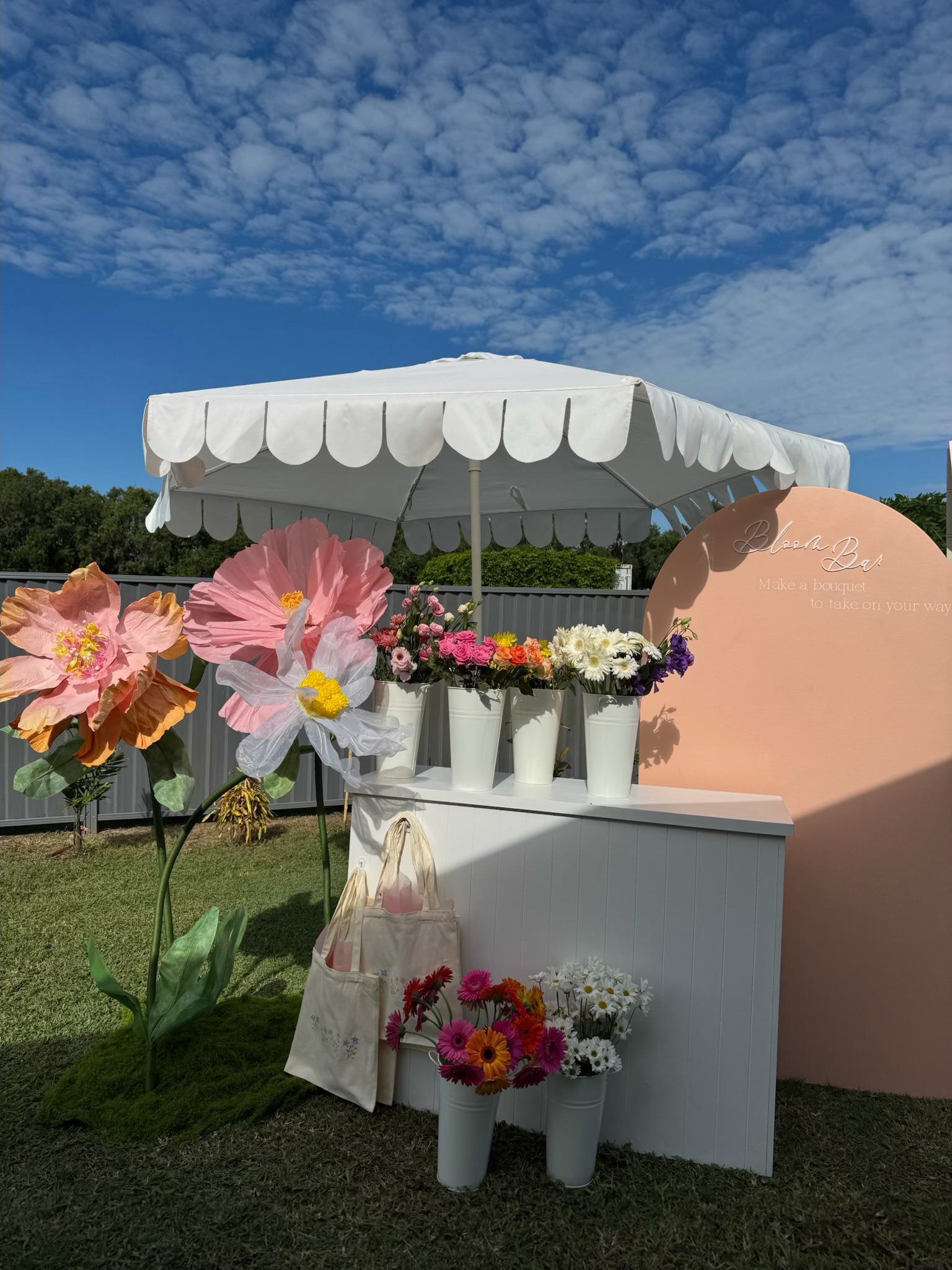A flower booth with white pots of colorful flowers under a white parasol, set against a pink backdrop with text, on a grassy area with a cloudy blue sky above.