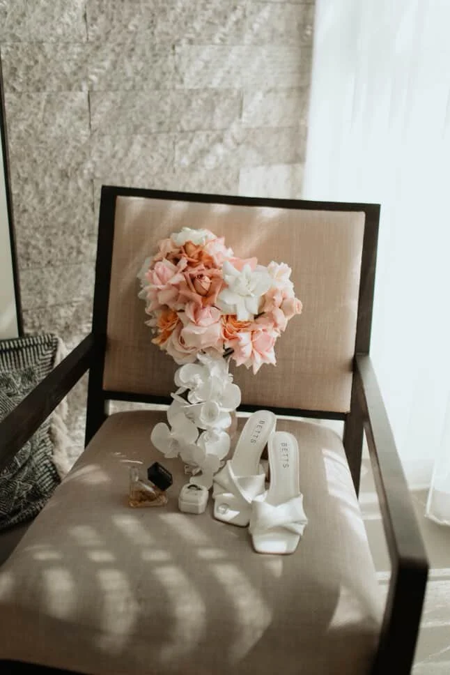 A beige upholstered chair with a dark wooden frame holding a pink and white flower arrangement, white high-heeled shoes, and small personal items on the seat near a sheer white curtain.