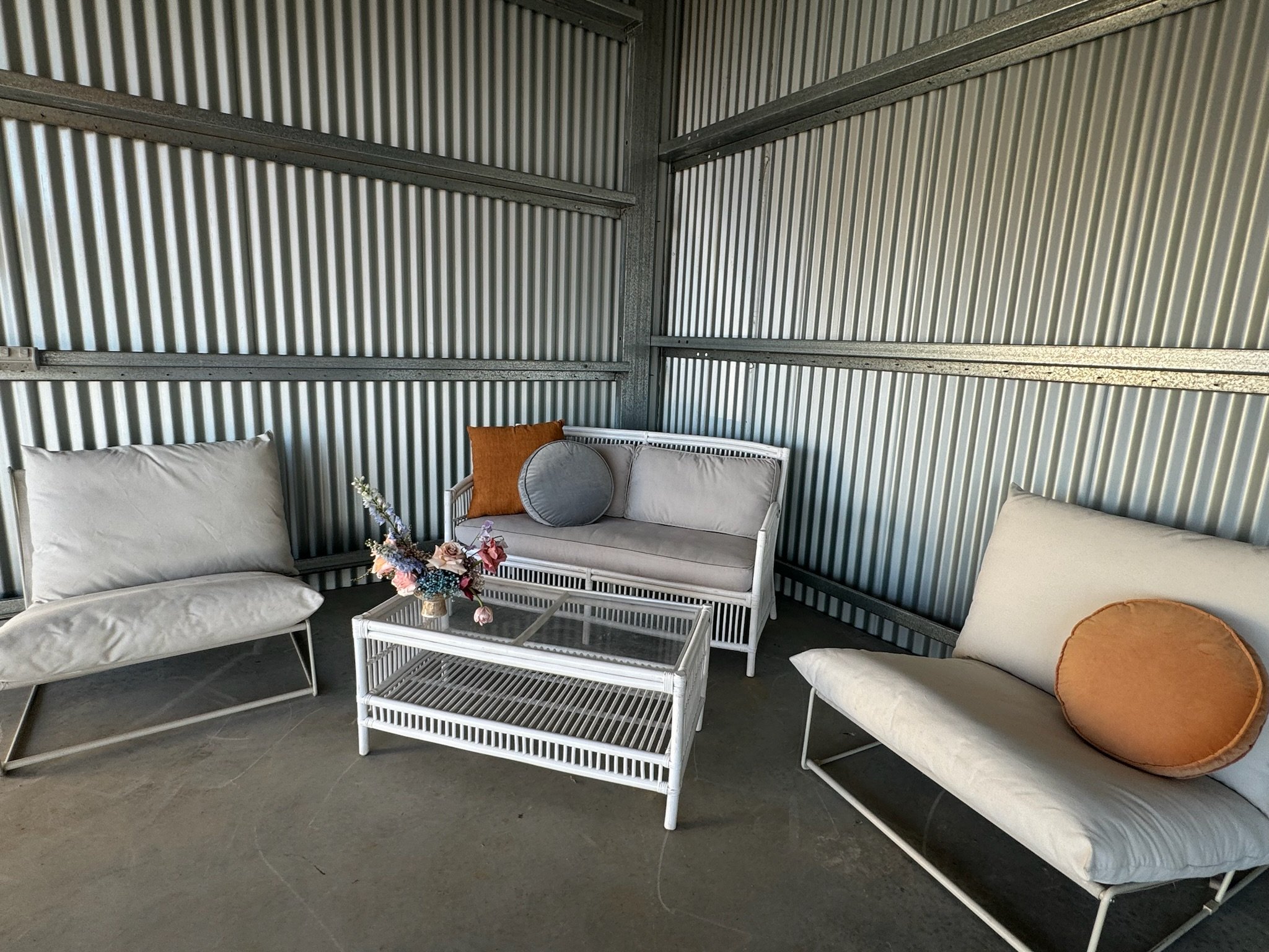 Interior of a metal shed with white outdoor furniture, including two lounge chairs with round and rectangular cushions, a glass-top coffee table with a flower arrangement, and a small white bench with cushions.