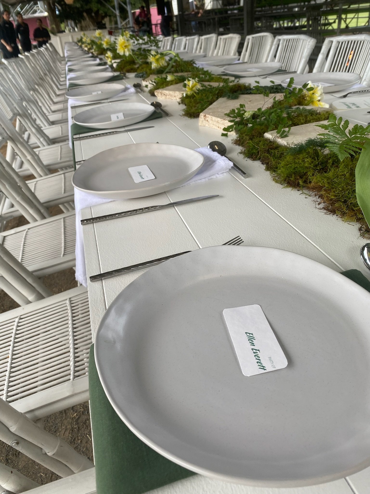 Long banquet table set outdoors with white plates, silver utensils, and name cards, decorated with greenery and flowers.