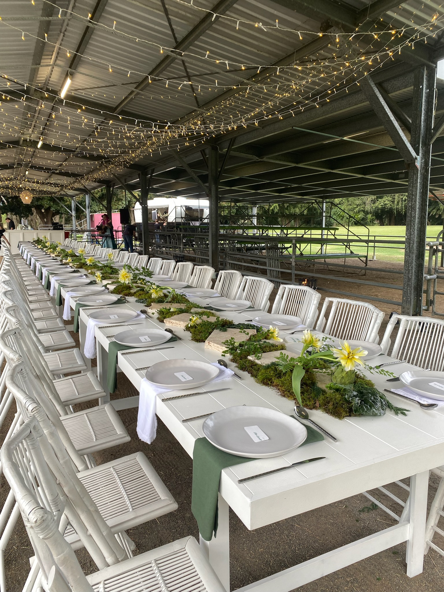 Long wedding reception table set under a metal tent with string lights. The table has white plates, silverware, green napkins, and a floral centerpiece with yellow flowers and greenery.
