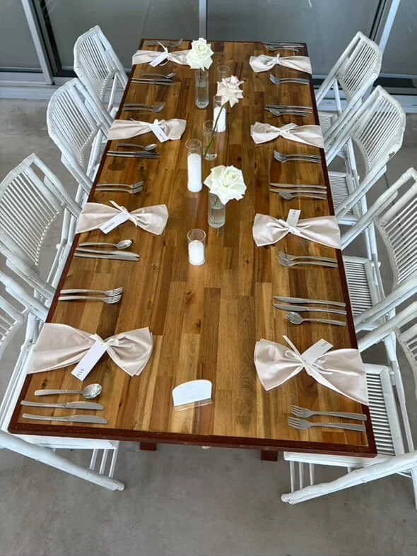 A rectangular dining table set with white chairs, beige bows as napkins, and minimalistic white flower centerpieces with candles.