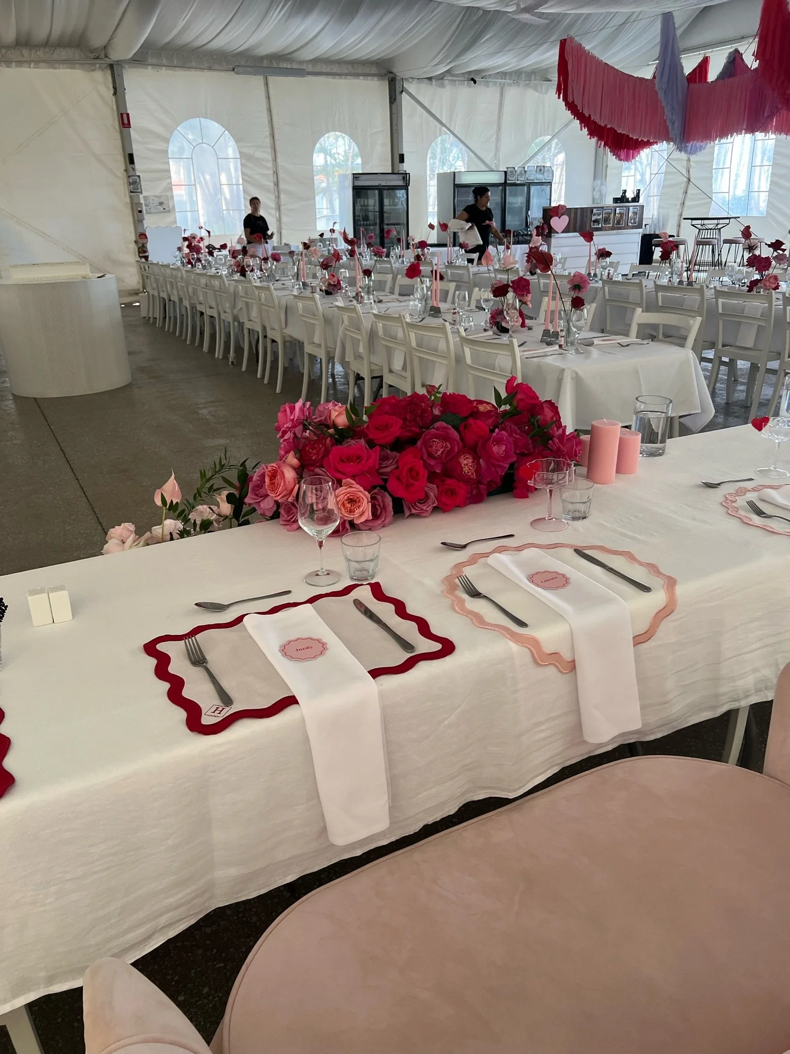 A wedding reception setup inside a large white tent with long tables, pink and red floral centerpieces, and pink candles. The tables are decorated with white tablecloths, pink placemats, and glassware, with some staff preparing the tables.