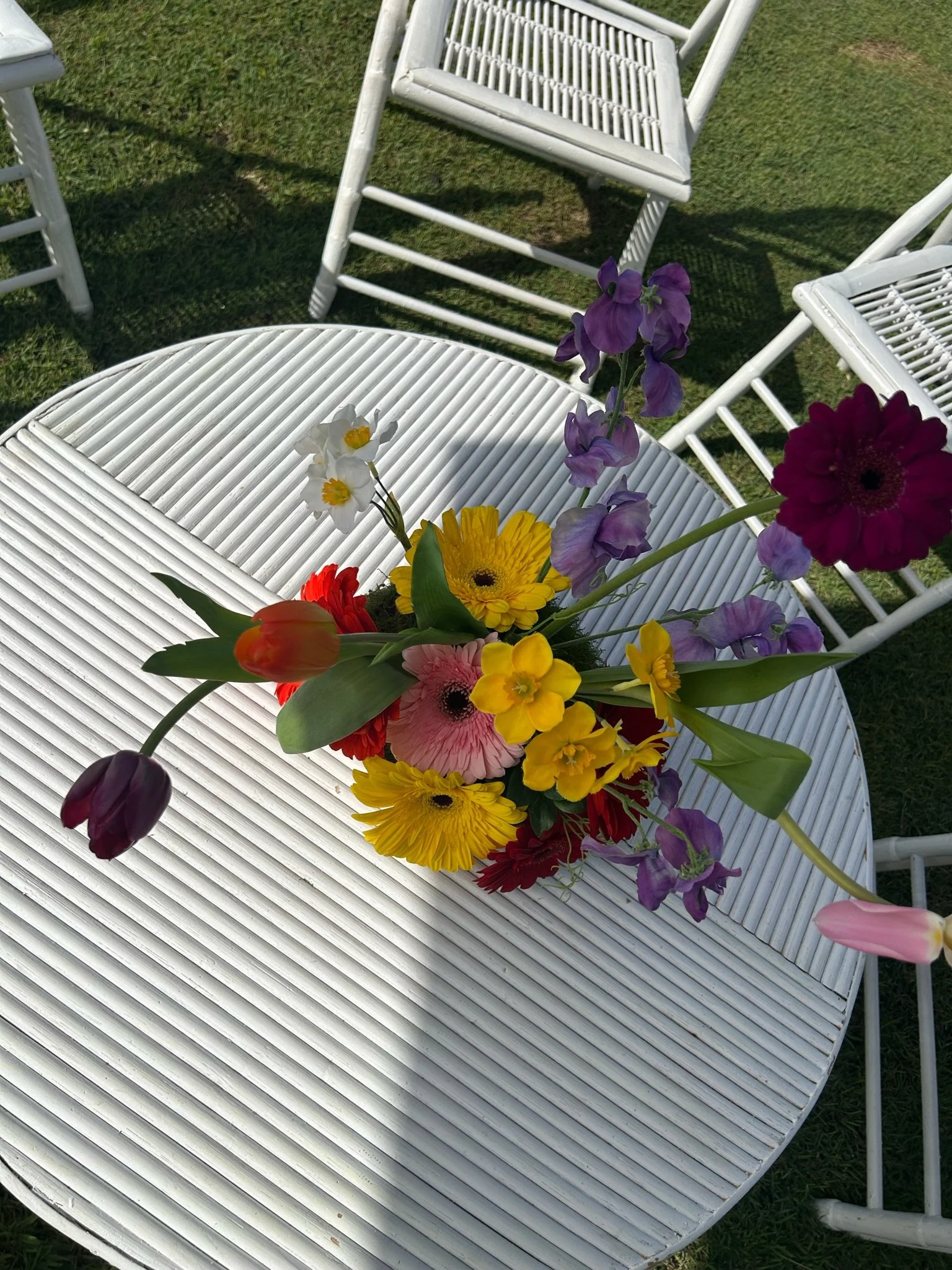 A colorful bouquet of flowers including tulips, daisies, and other varieties on a white outdoor table with white chairs and green grass in the background.