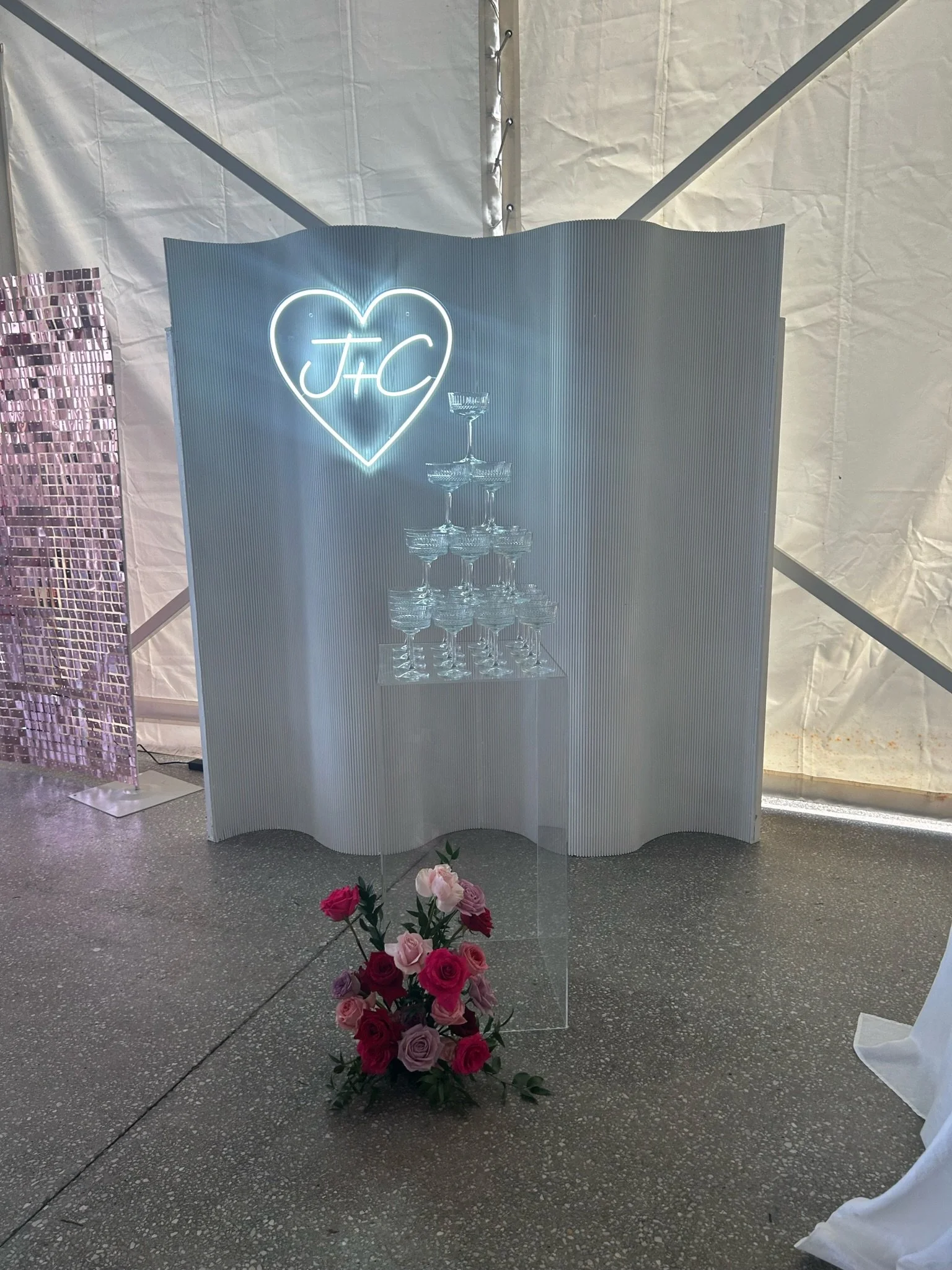 Neon sign with a heart and initials J and C, a champagne tower made of glasses, and a bouquet of pink and purple roses in front of a white decorative backdrop.