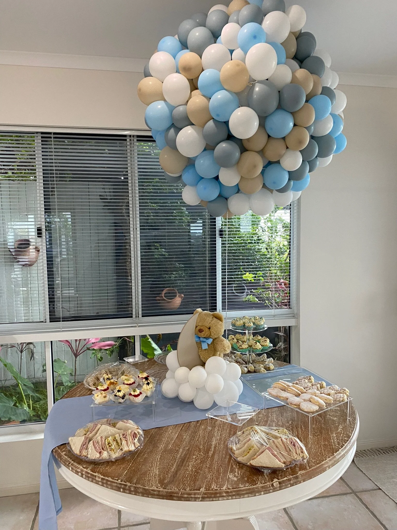 A teddy bear wearing a blue bow tie on a cloud-like arrangement of white balloons, with a large balloon cluster above. The setup is on a wooden table with glass jars of desserts, sandwiches, and cupcakes, in front of a window with blinds and greenery