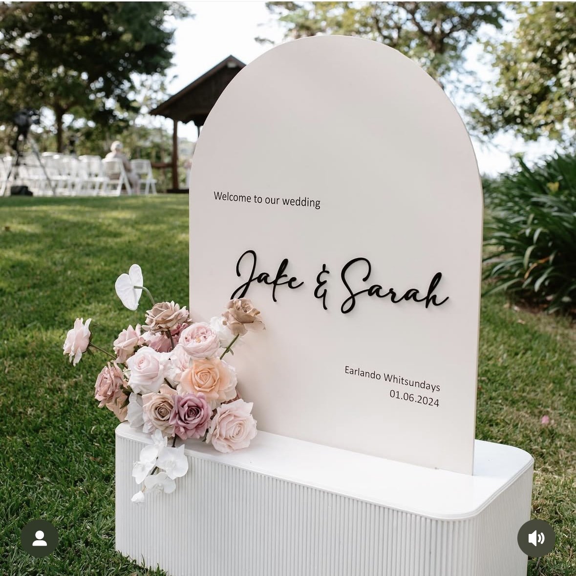 Wedding welcome sign with pink, purple, and white flowers, set outdoors with chairs in the background.