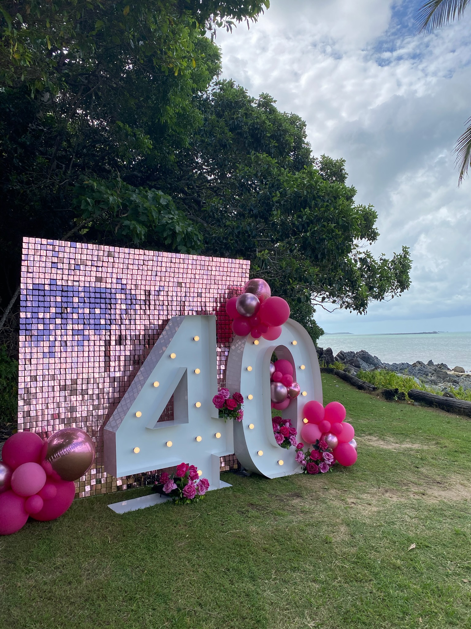 Large white illuminated number 40 decorated with pink and metallic balloons and pink roses, positioned outdoors on green grass with a background of trees, water, rocks, and partly cloudy sky.