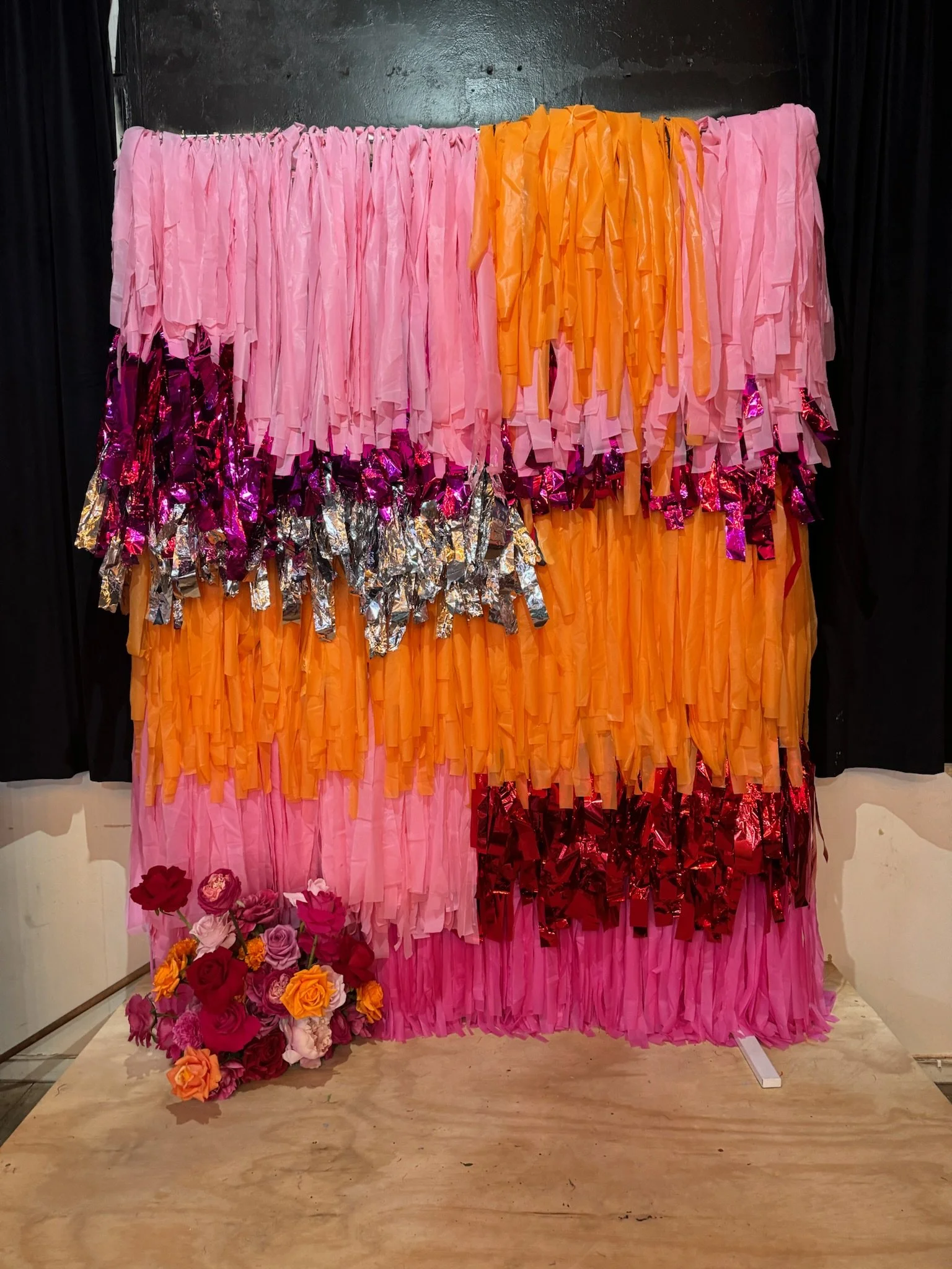 Colorful decorative backdrop with pink, orange, purple, silver, and red fringed paper or fabric strips, with a bouquet of pink, orange, and purple flowers at the bottom left corner.