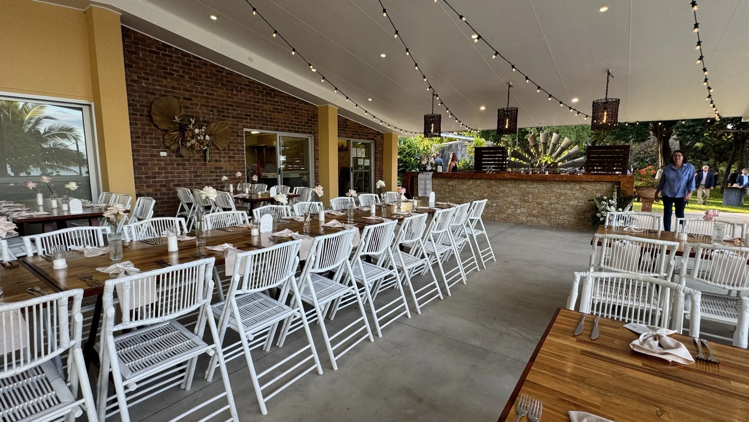 Set up for a celebration or event with long dining tables decorated with white tablecloths, vases with flowers, and napkins, in an outdoor covered patio area with brick and yellow painted walls, string lights, and a bar in the background. People are 