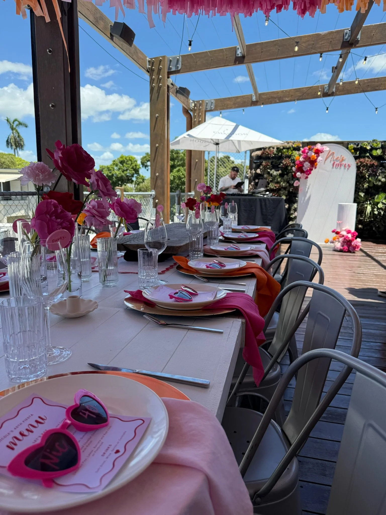 Decorated outdoor dining table with pink and orange napkins, plates, glasses, and pink flower centerpieces under string lights, with a DJ in the background and floral backdrop with 'Mommy' sign.