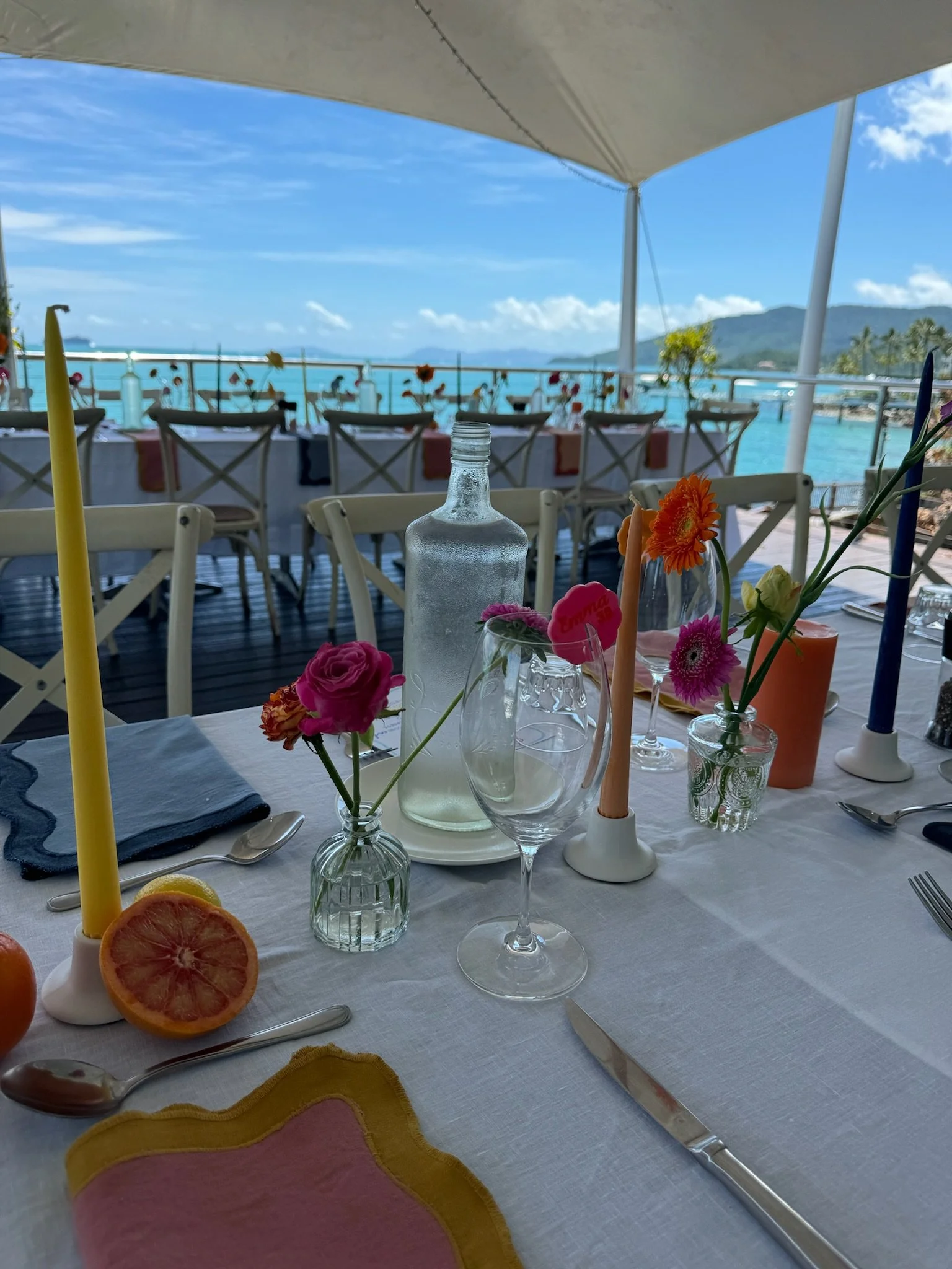 Decorated table set for a meal on a patio overlooking a body of water and mountains, with colorful flowers, candles, water bottles, and glassware.