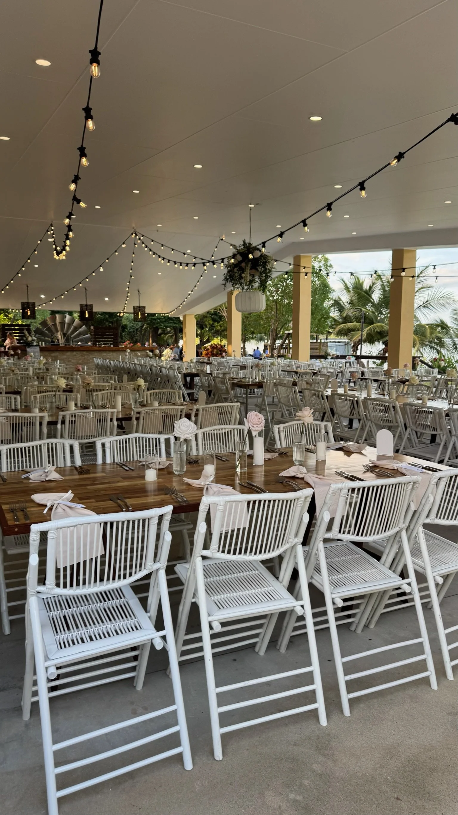 Wedding or event reception setup with wooden tables, white chairs, candles, flowers, and string lights under a covered patio with view of palm trees.