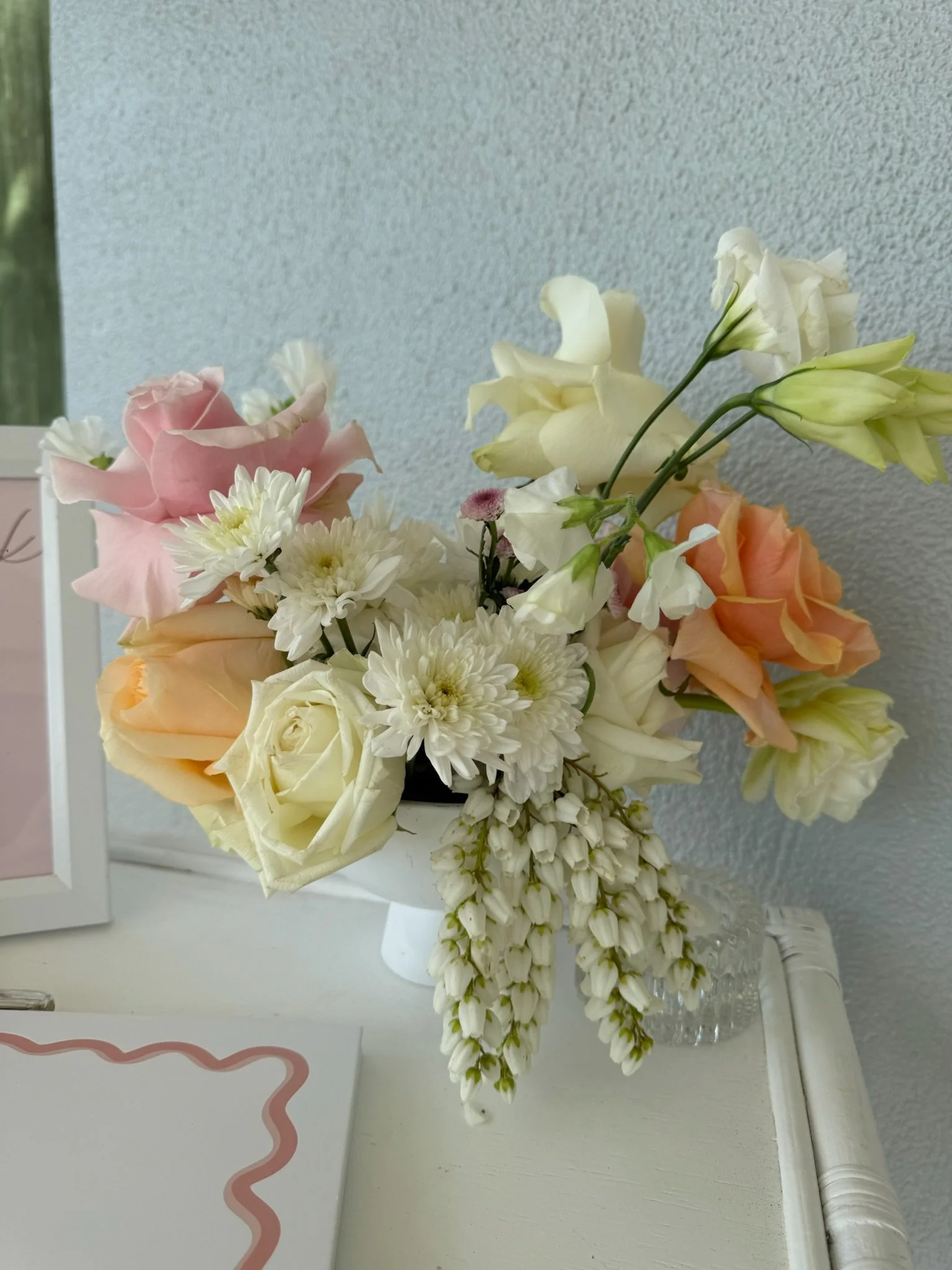 A bouquet of white, pink, peach, and cream-colored flowers in a glass vase on a white surface against a light-colored textured wall.