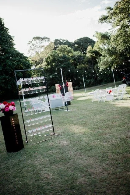 Outdoor event setup with white chairs, tables, and string lights on a grassy area surrounded by trees.