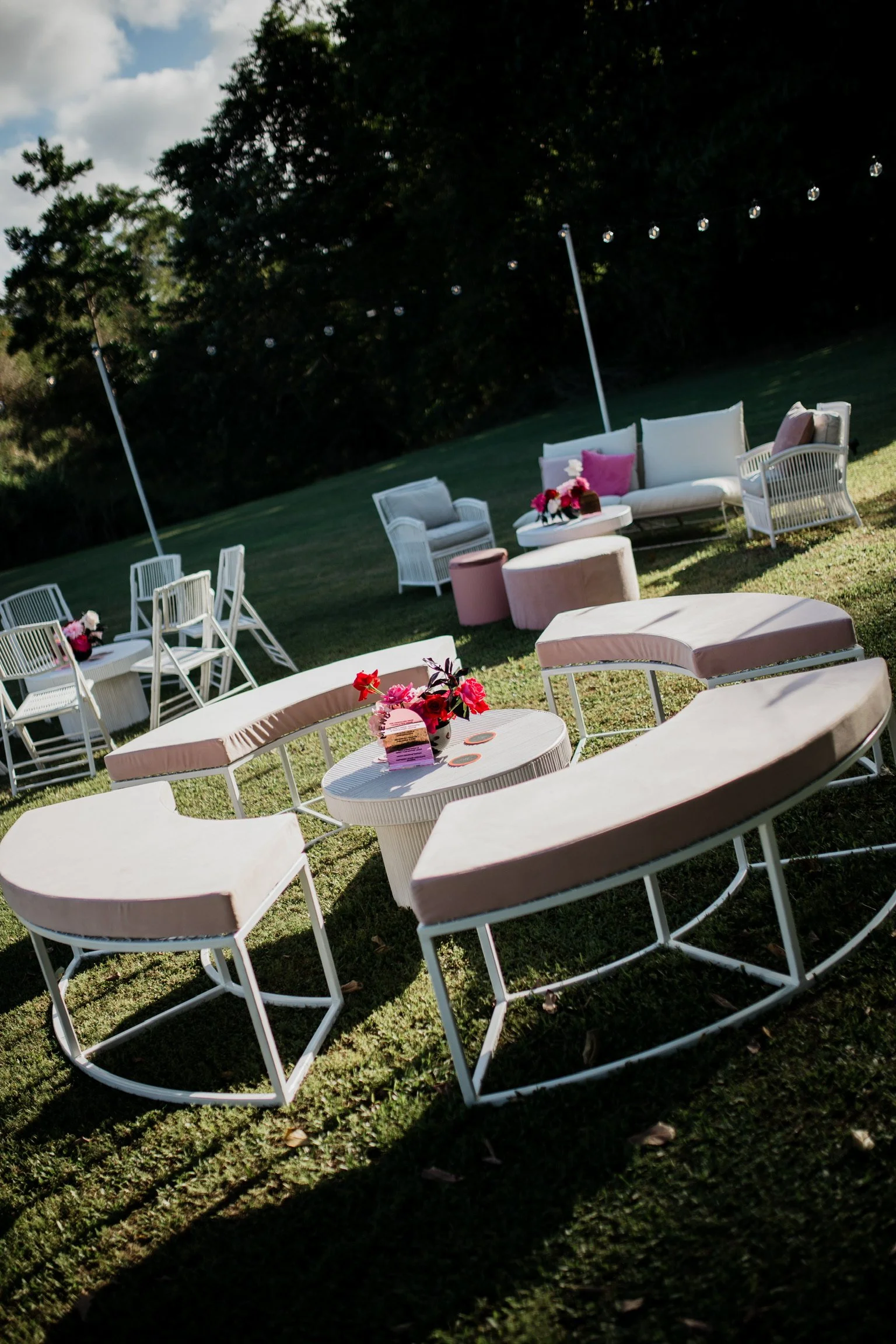 Outdoor lounge area with white cushioned chairs, pink and red flower arrangements on tables, string lights overhead, and a grassy lawn with trees in the background.