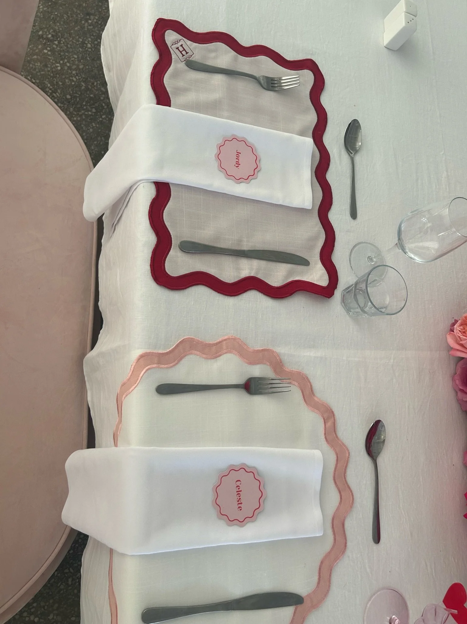 Table setting with two place settings, each with a white napkin, a fork, knife, and spoon, decorated with pink and red scalloped placemats, with name tags labeled 'Judy' and 'Celeste'.