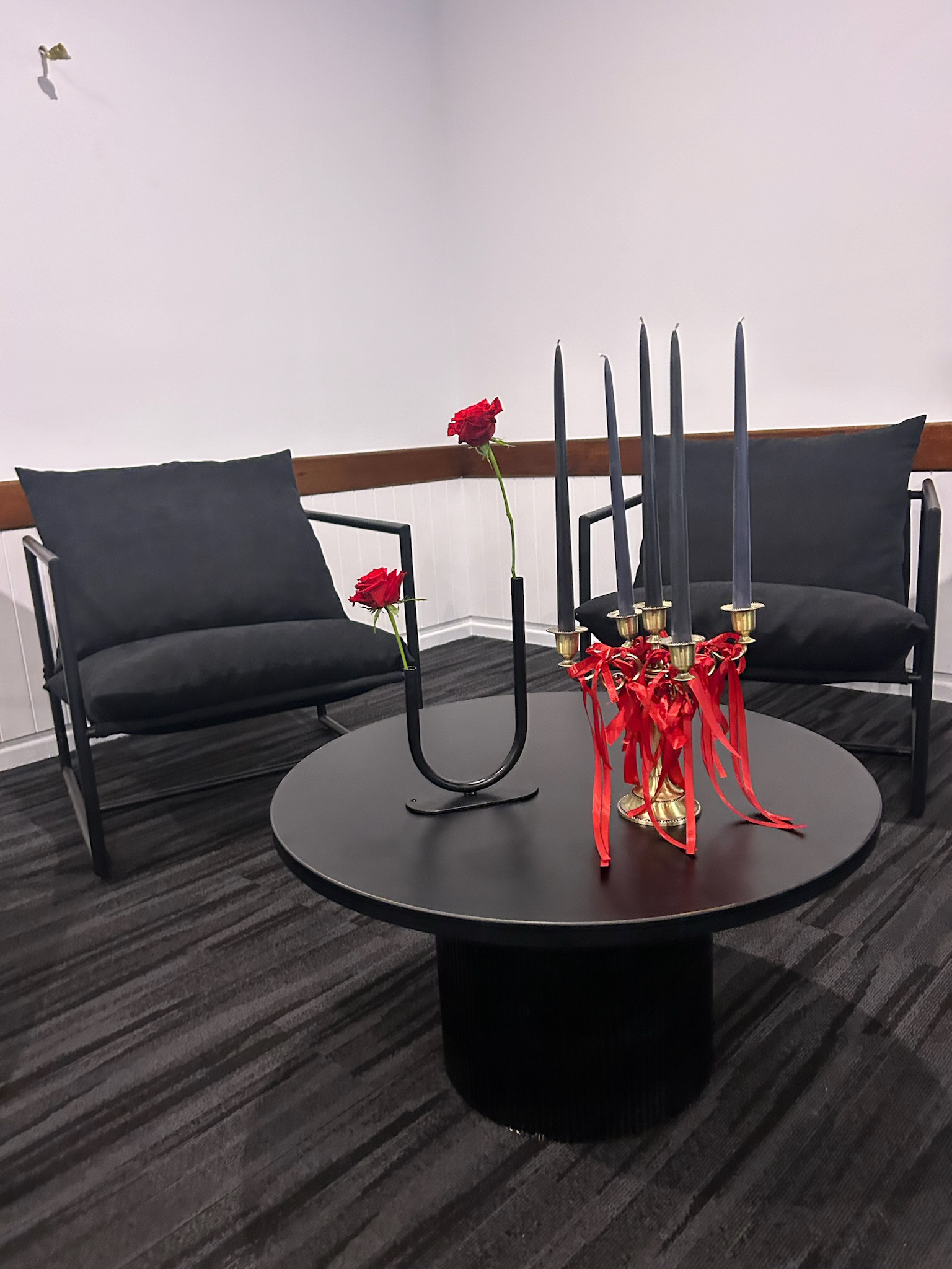 Two black chairs with dark cushions facing a round black table with a golden candelabrum with five black candles and red ribbons, and two red roses on the table.