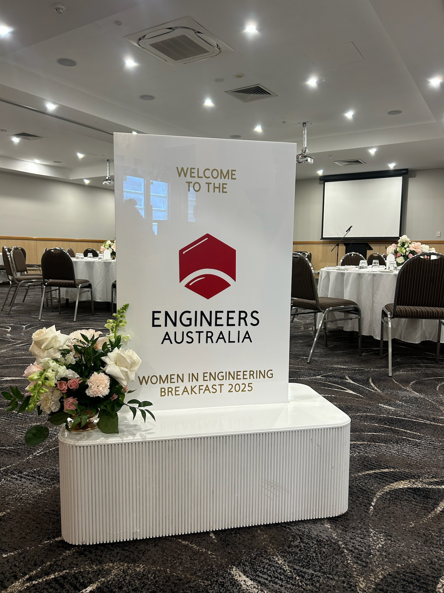 Sign for the Engineers Australia Women in Engineering Breakfast 2025 event, with floral arrangement in a white container in the foreground, in a conference room with round tables and chairs, and a projector screen in the background.