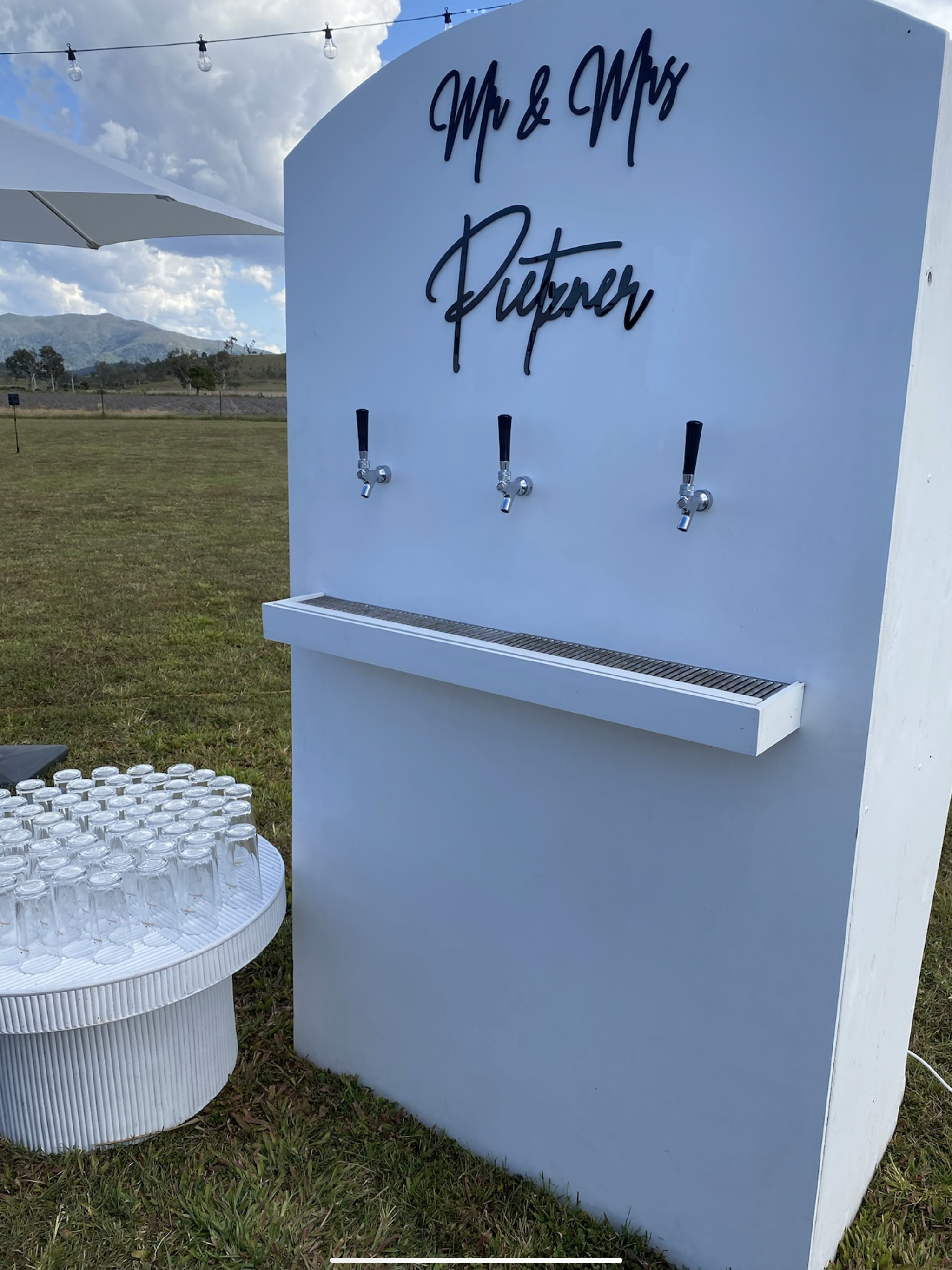 White outdoor drink dispenser with three taps, labeled 'Mr & Mrs' and 'Pitter' for wedding or event. There are glasses on a nearby table, and the scene is set in a grassy outdoor area with mountains and cloudy sky in the background.