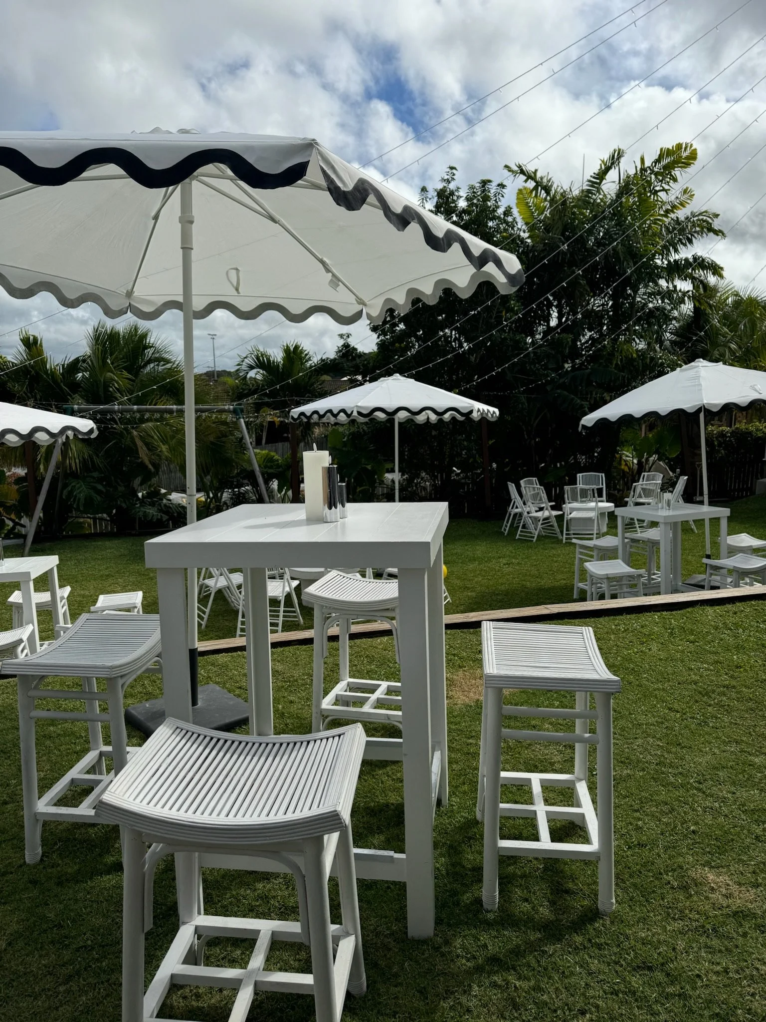 Outdoor patio with white high-top tables and chairs, large white umbrellas, green grass, palm trees, cloudy sky, and electrical wires overhead.