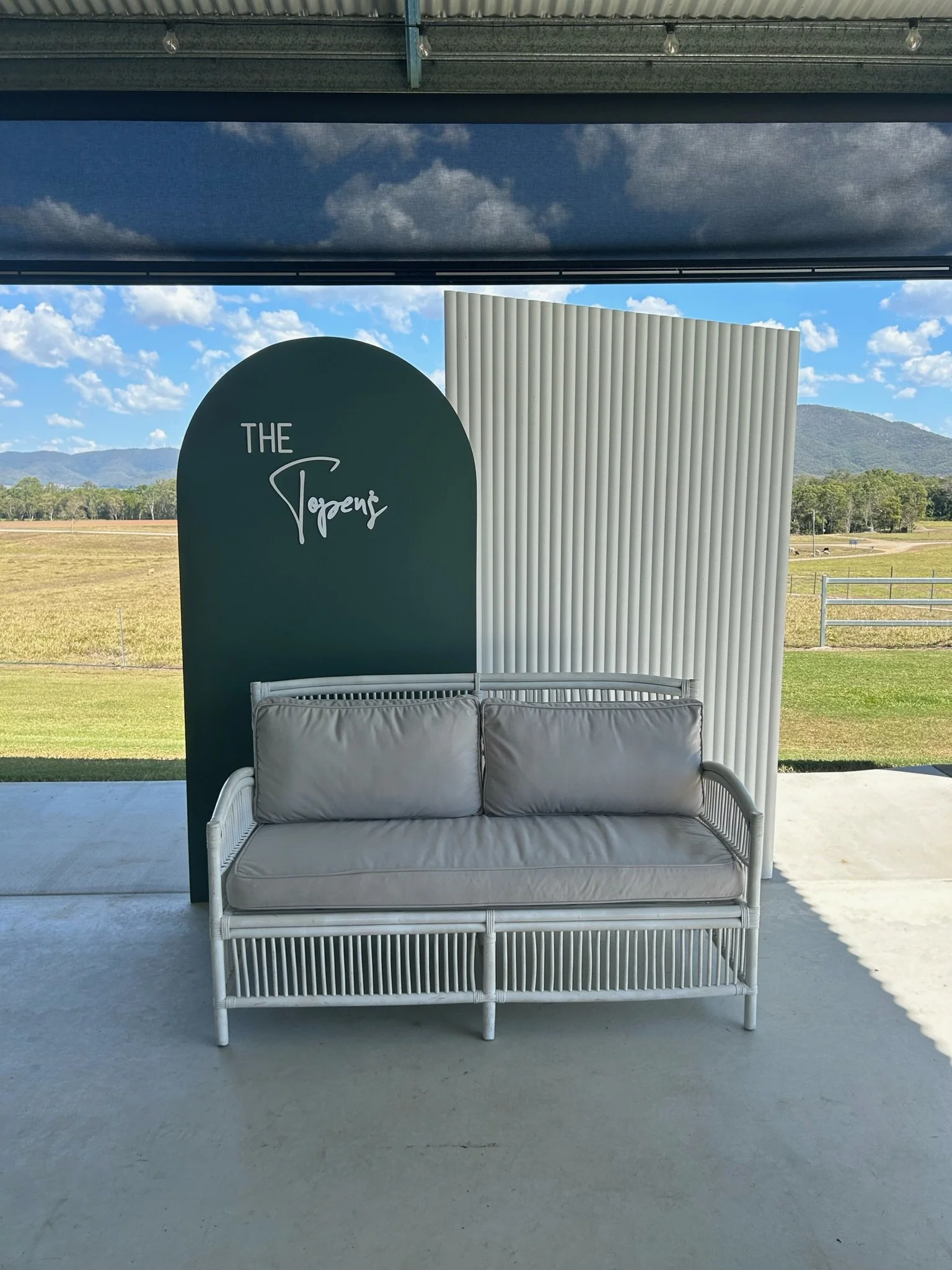 An outdoor patio area with a white wicker sofa with beige cushions, positioned in front of two decorative panels, one dark green with white text that reads 'The Trogans' and the other white with vertical ridges, against a scenic background of fields,