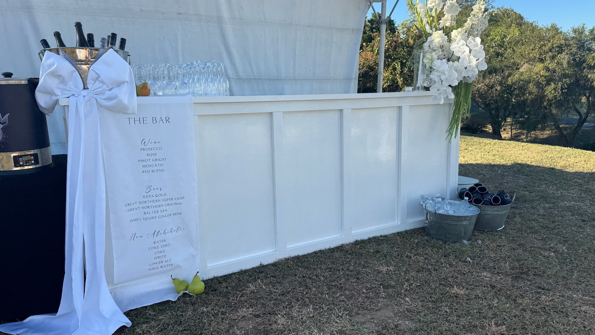 Outdoor beverage station with a white bar, flower arrangement, and buckets of drinks and cups at a garden event.