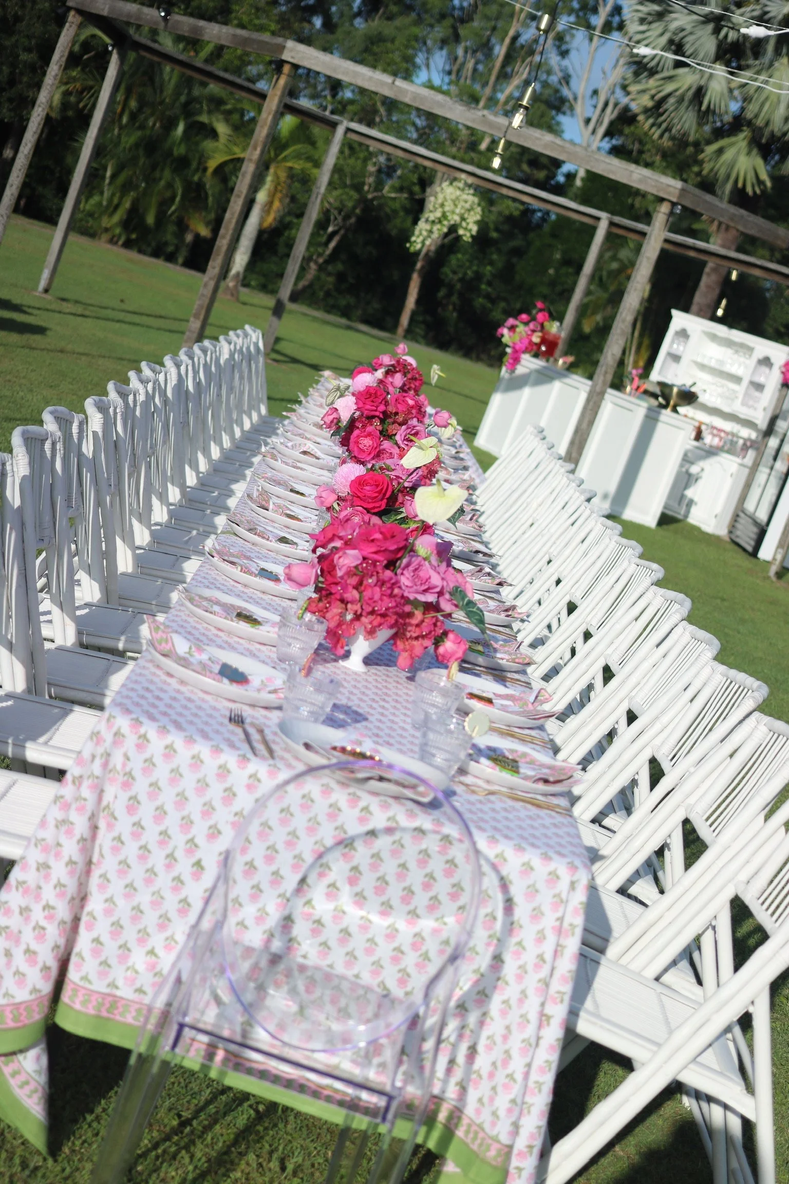 Outdoor event setup with a long table decorated with pink flowers, white chairs, and place settings, in a grassy area under trees with string lights overhead.